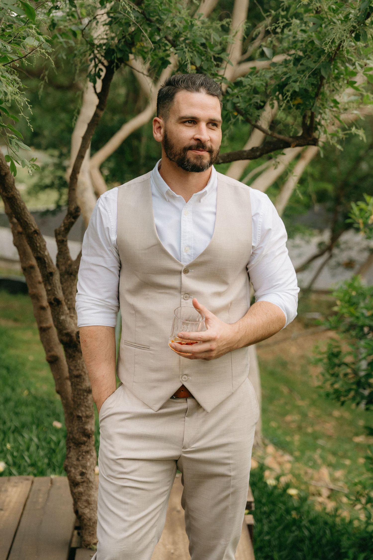 Person in beige vest and white shirt holds a drink in an outdoor garden setting.