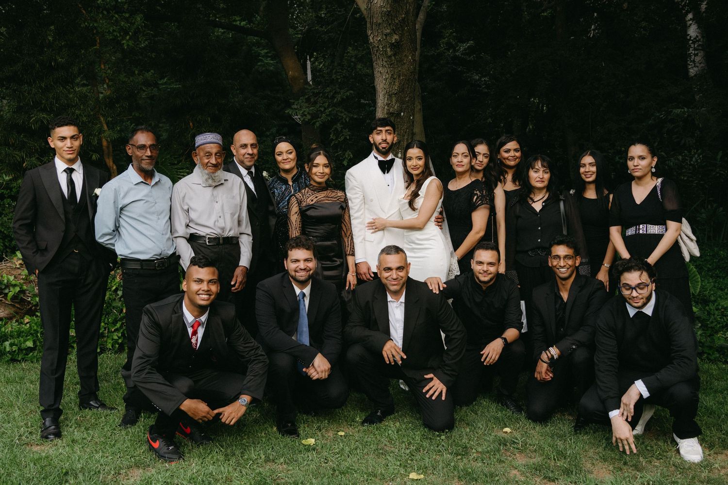 Large wedding party group photo with guests in formal attire posed outdoors.