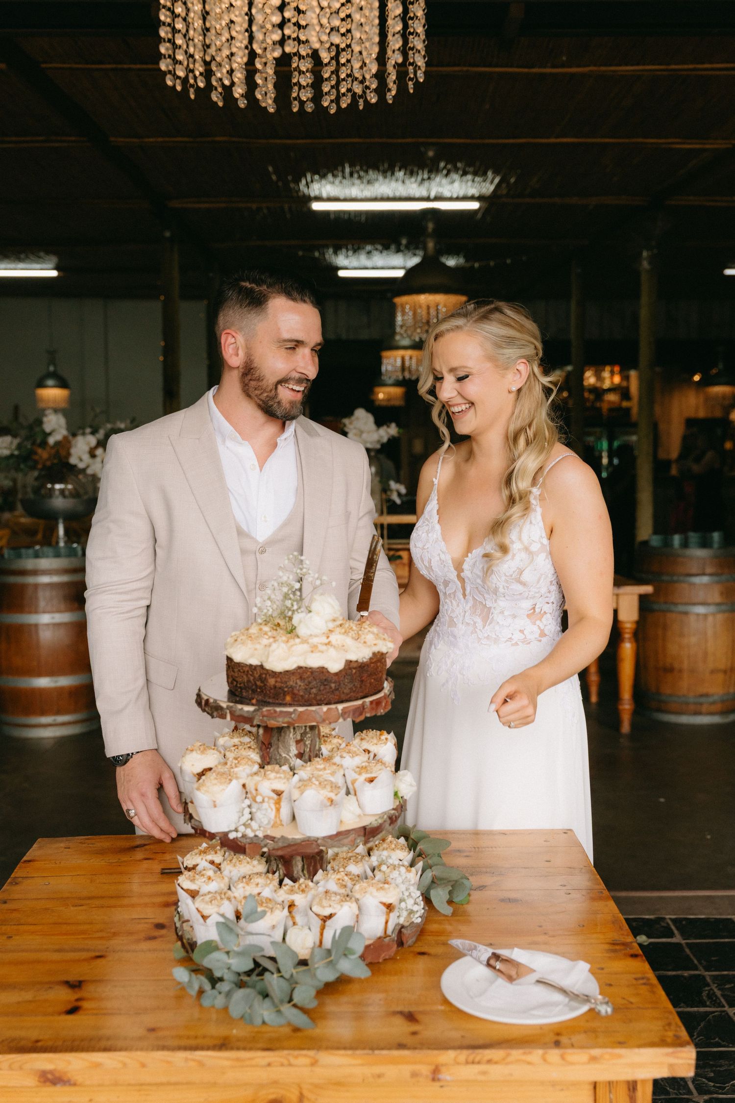 A couple cuts their rustic wedding cake decorated with flowers at a wooden table.