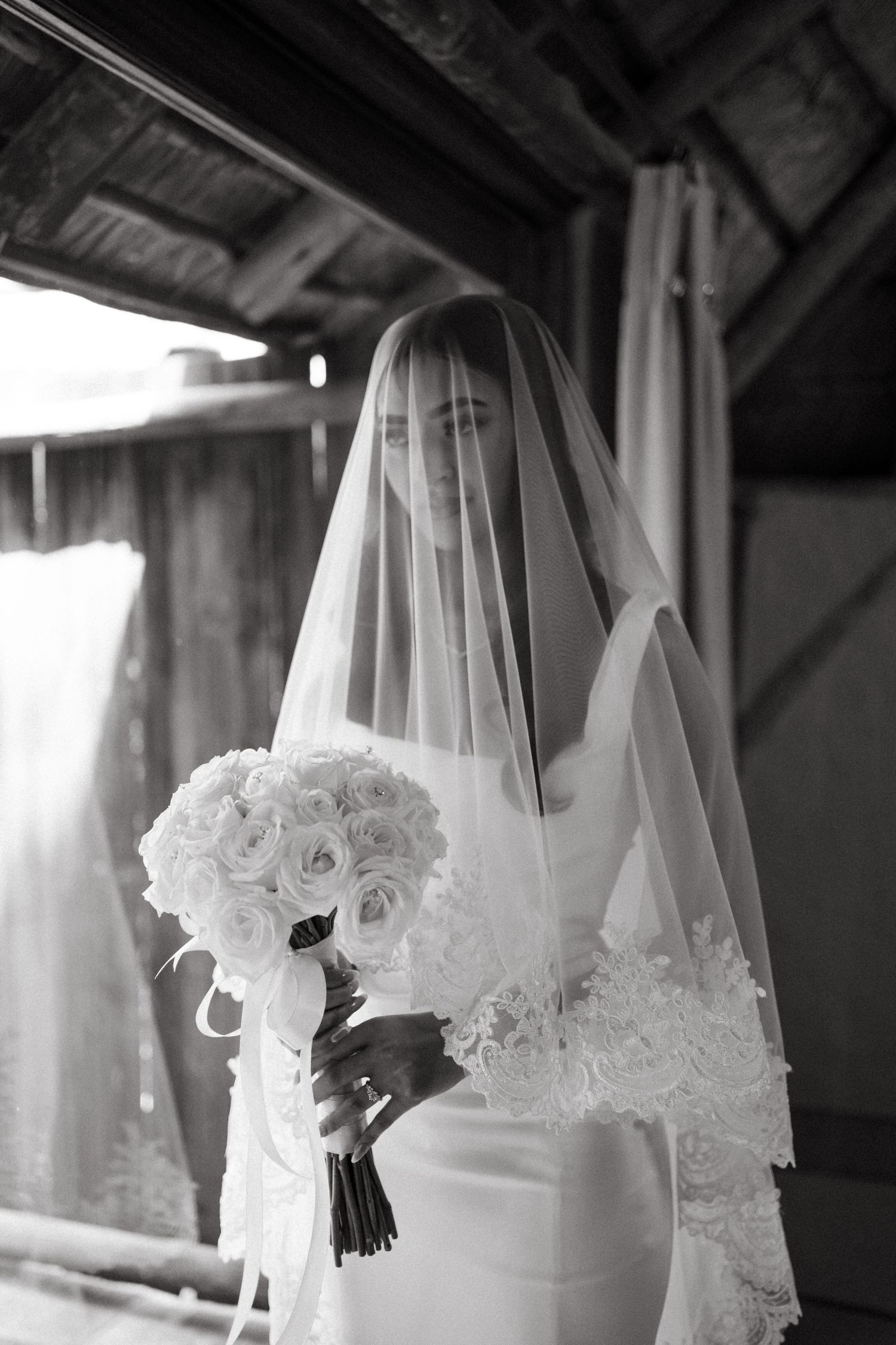 An elegant bride in a white veil holds a bouquet of white roses in a moody black and white photograph.