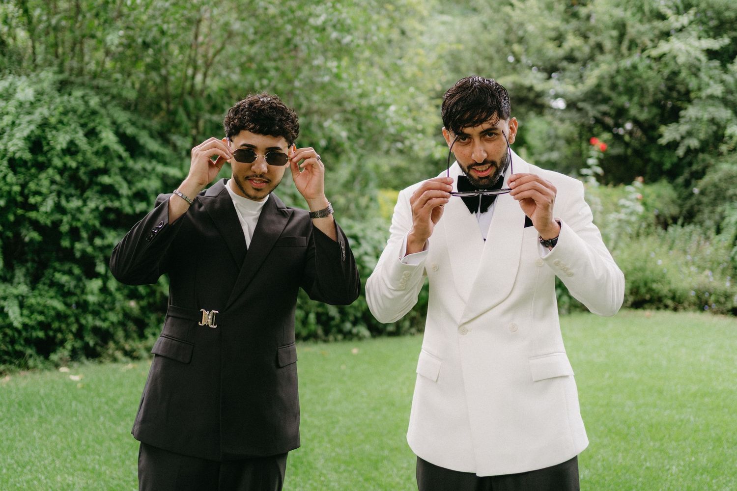 Two friends in formal attire adjusting their sunglasses while standing on grass with greenery in background.