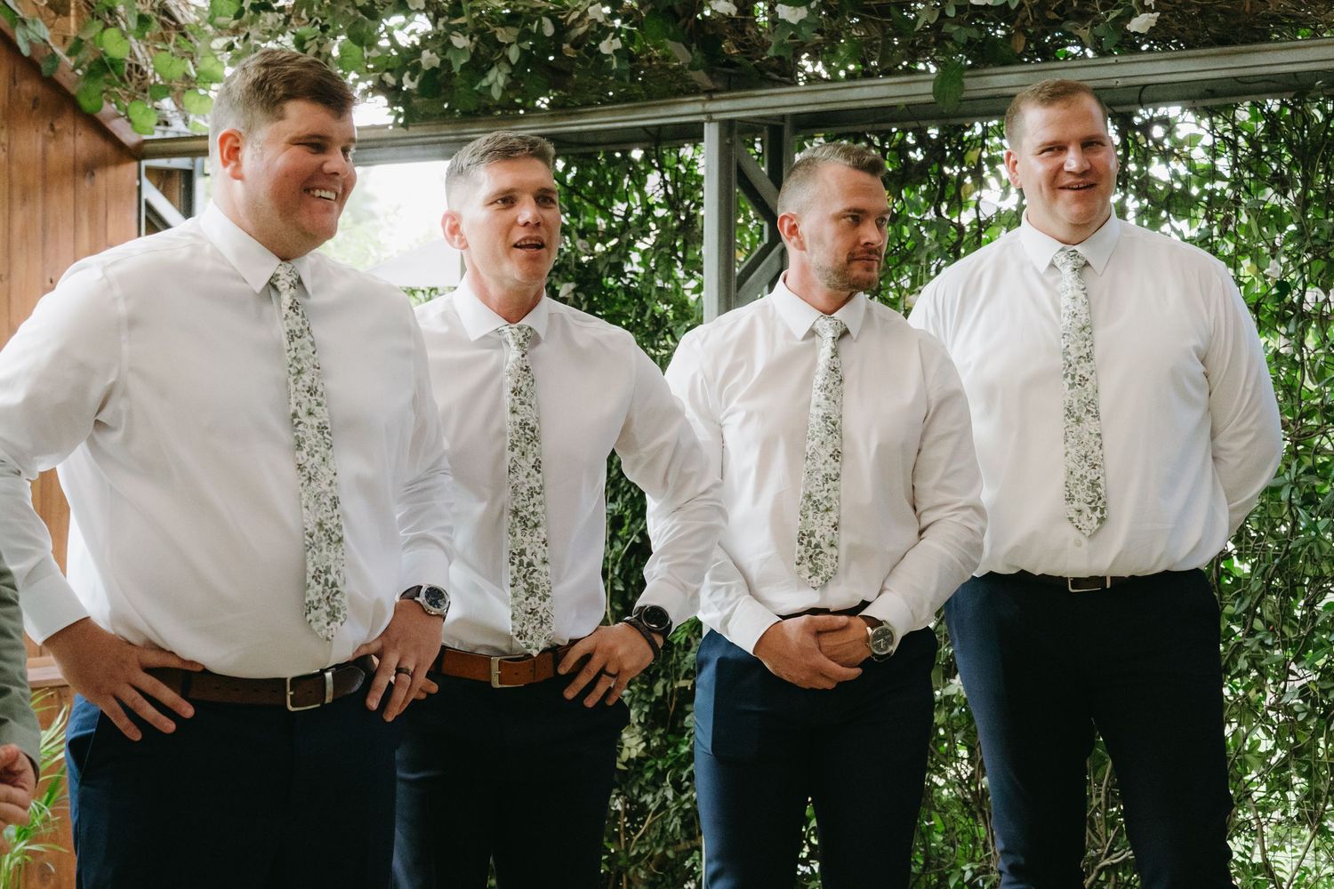 Four groomsmen in white shirts and floral ties pose together outdoors against greenery.