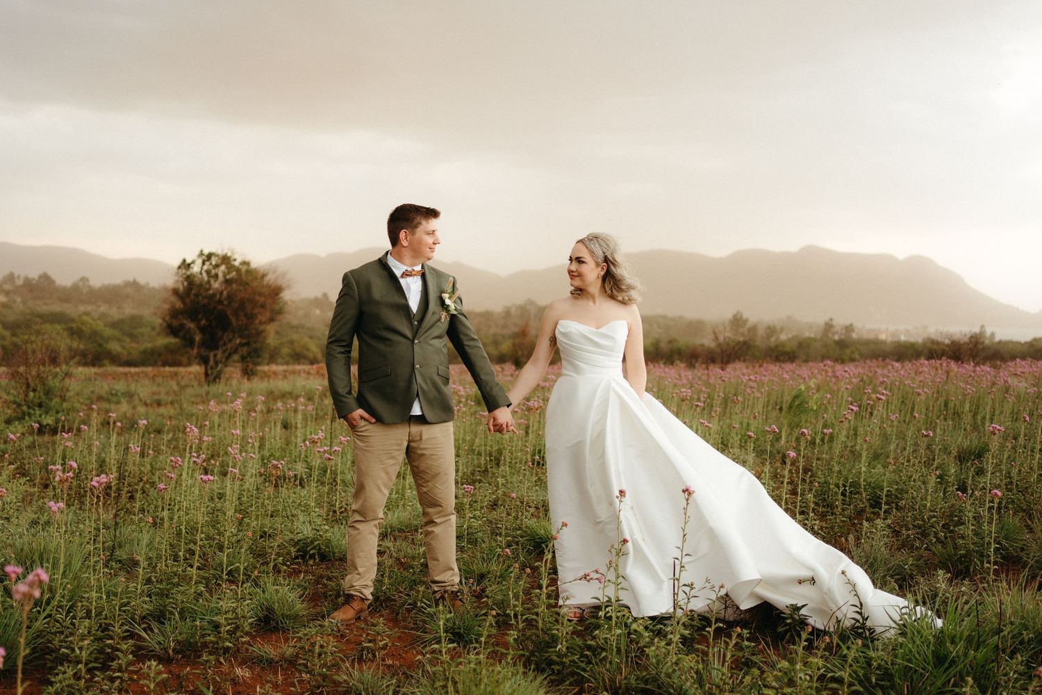 A couple walks hand in hand through a misty mountain meadow at sunset, with the bride's white dress flowing in the breeze.
