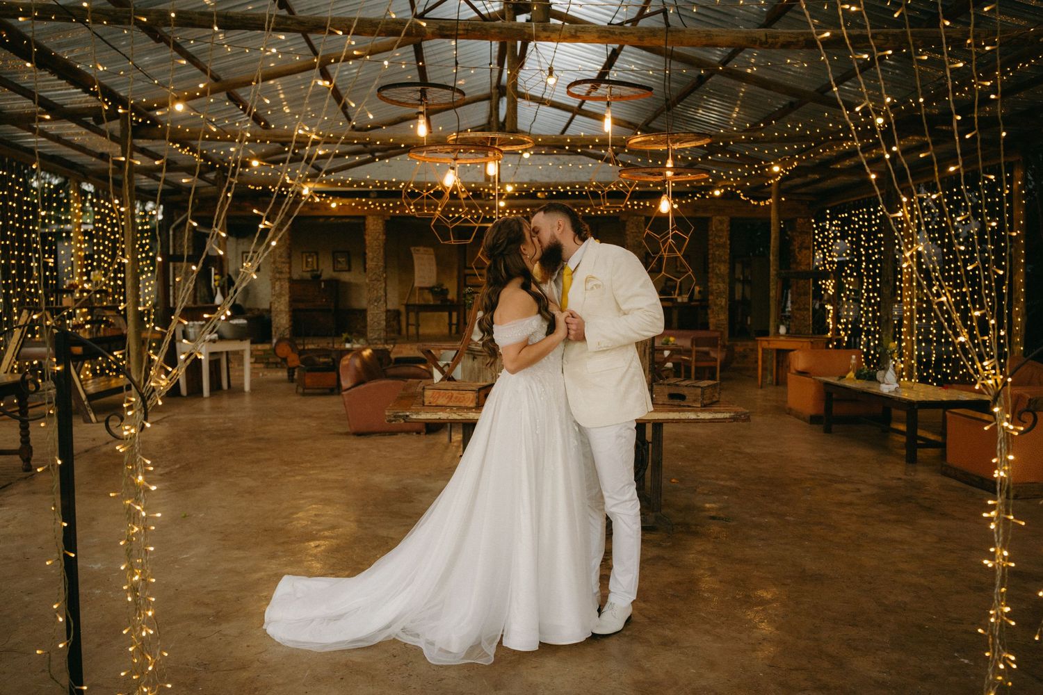 A wedding couple dances in a rustic barn venue decorated with twinkling string lights.