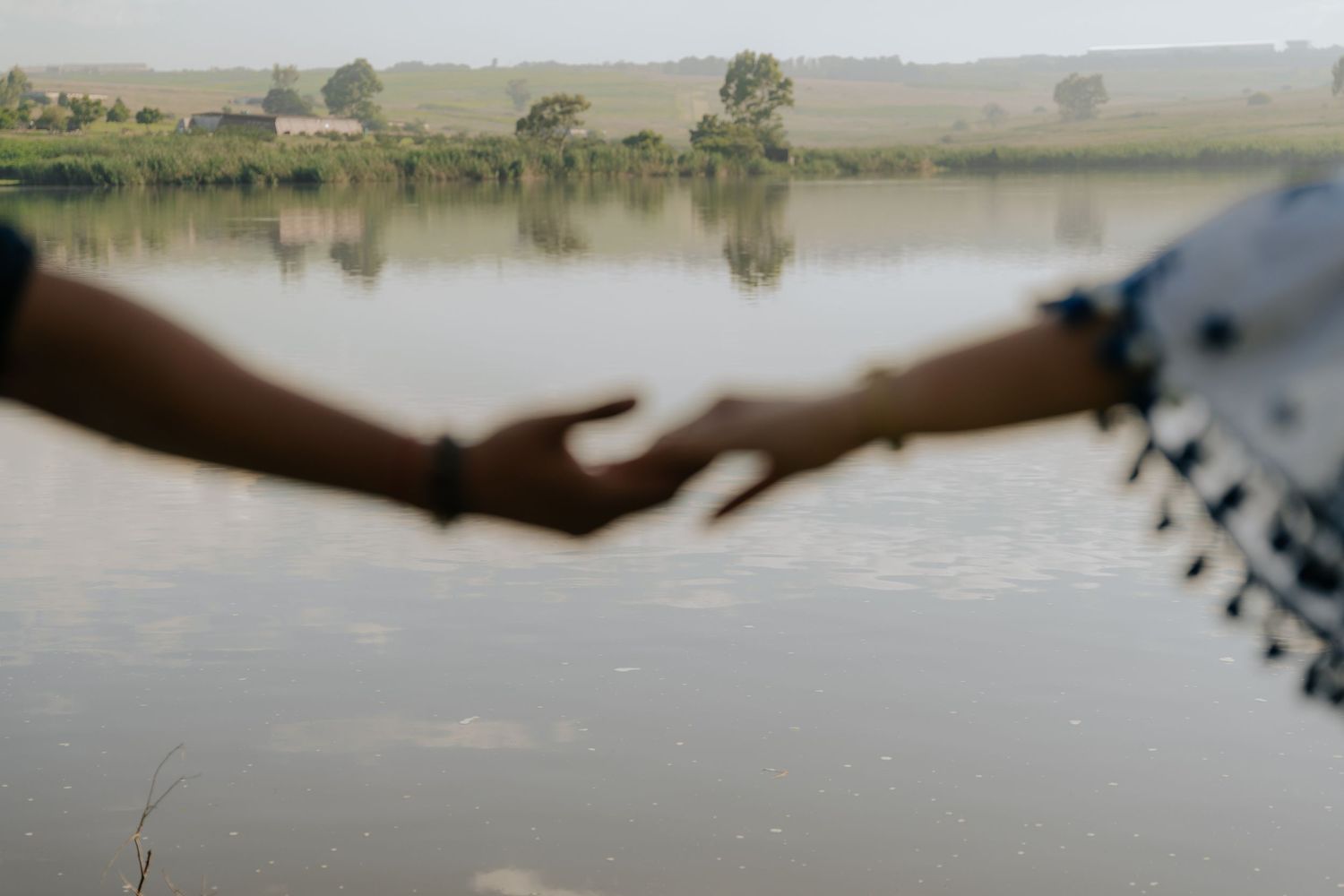 Two hands reach out towards each other across a tranquil lake with trees and reflections in the background.