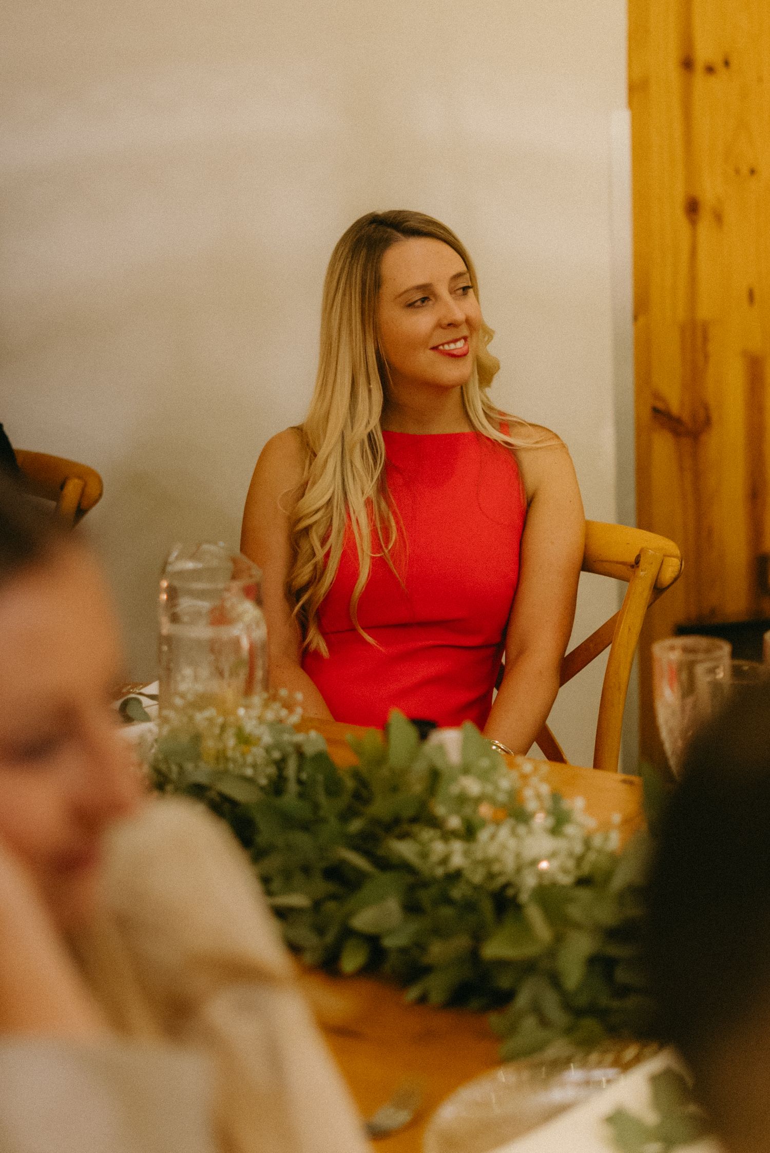 A person in a red sleeveless dress sits at a wedding reception table decorated with greenery.