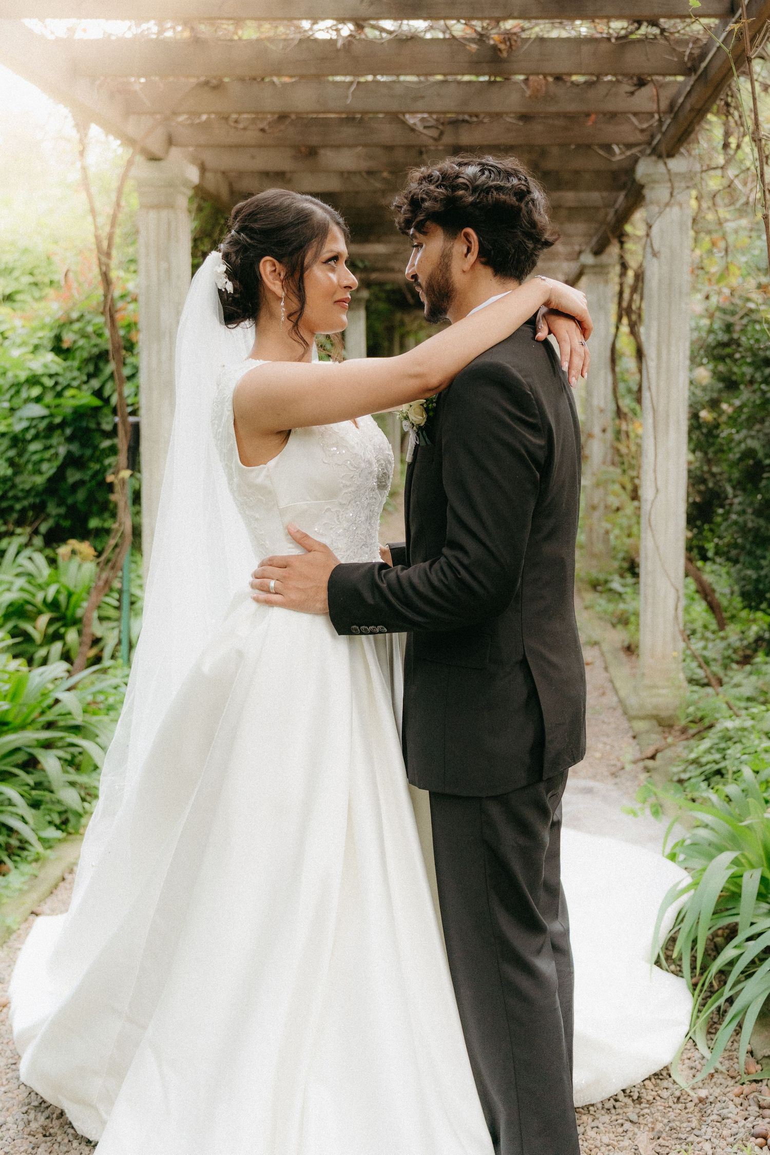 A romantic wedding couple embracing under a pergola in a garden setting.