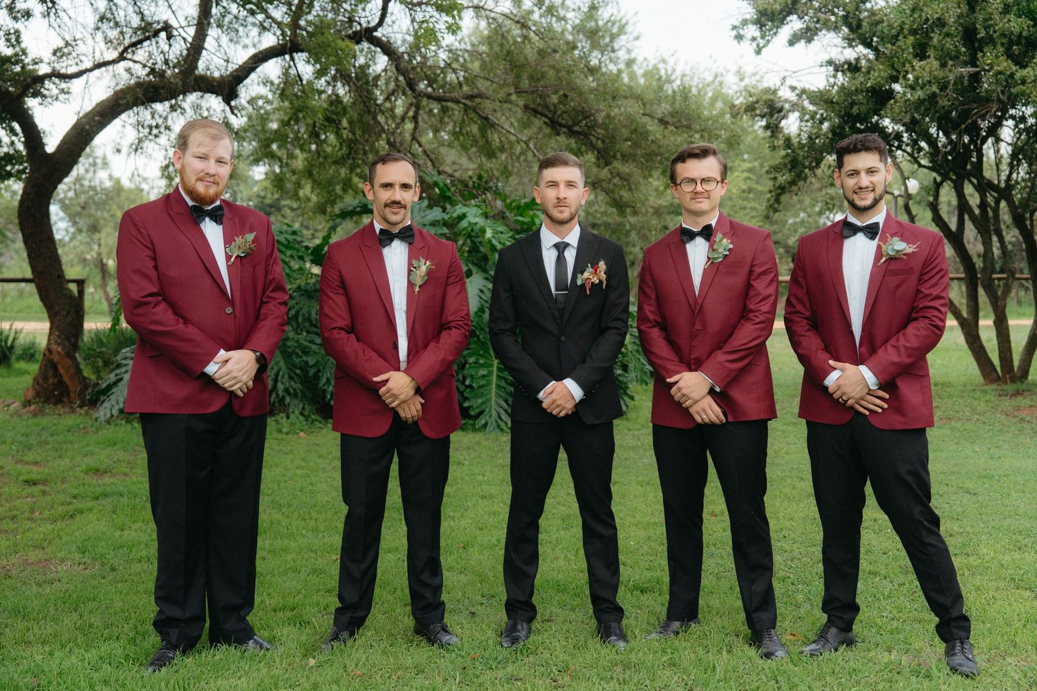 Wedding party group in coordinated burgundy tuxedo jackets and black bow ties standing outdoors.