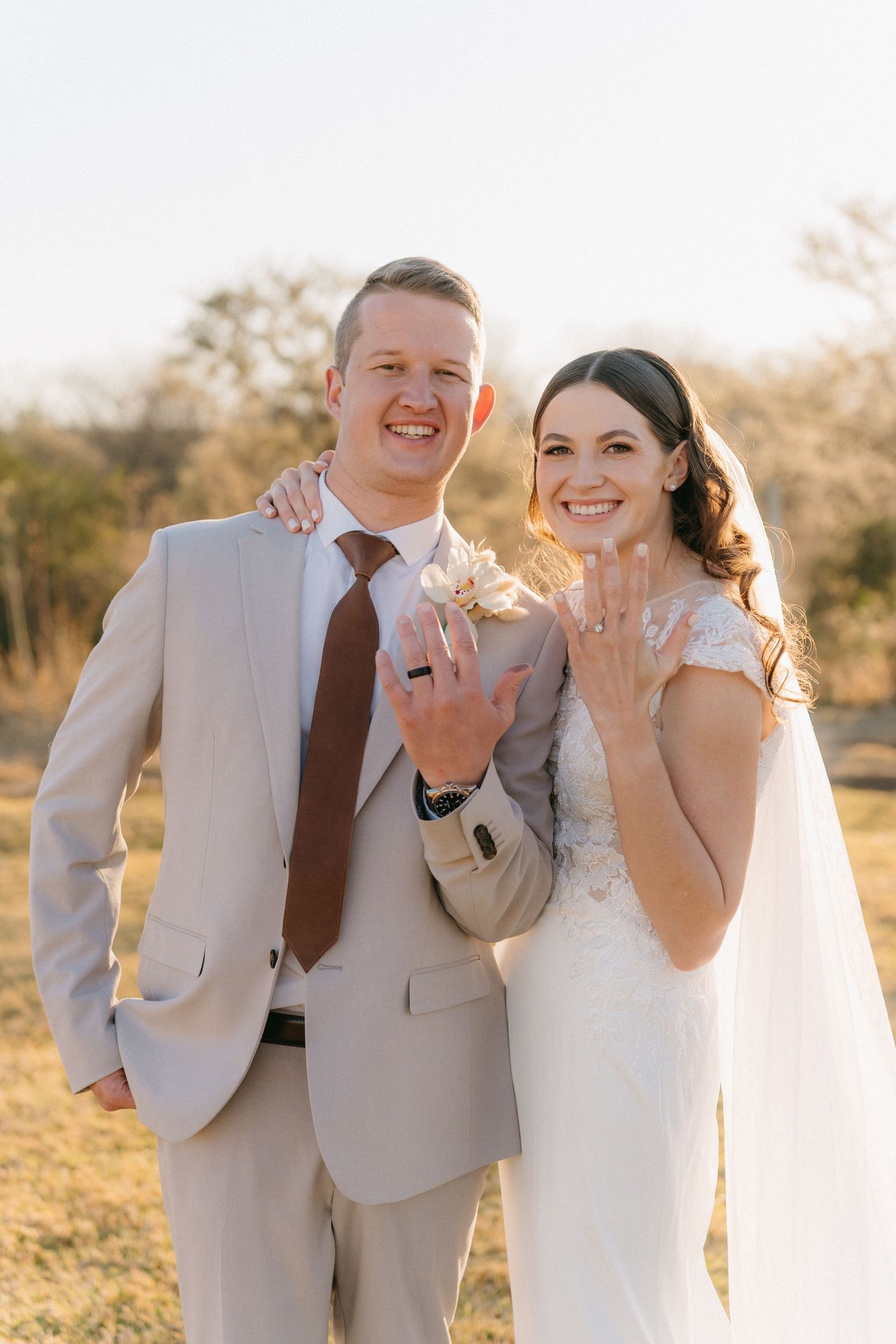 A happy couple poses in formal wedding attire during golden hour in a natural outdoor setting.