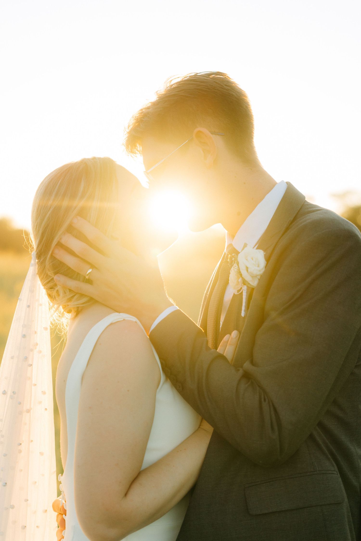 A silhouetted couple embraces tenderly in golden sunset light with a bride in white dress and groom in dark suit.