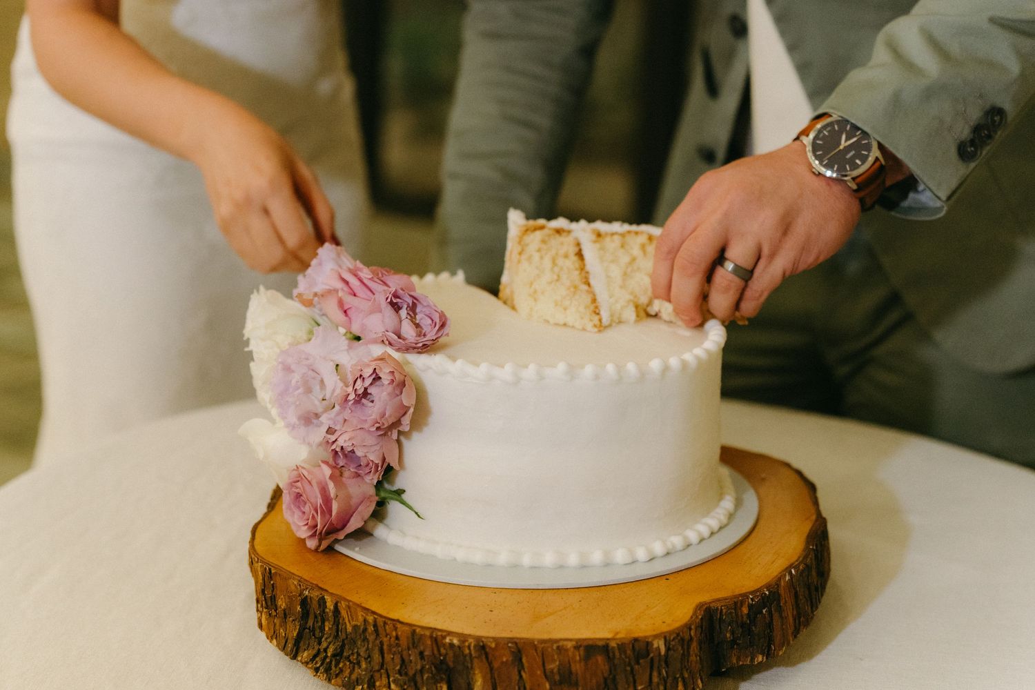 Wedding cake with pink floral decorations being cut on rustic wood slice stand.