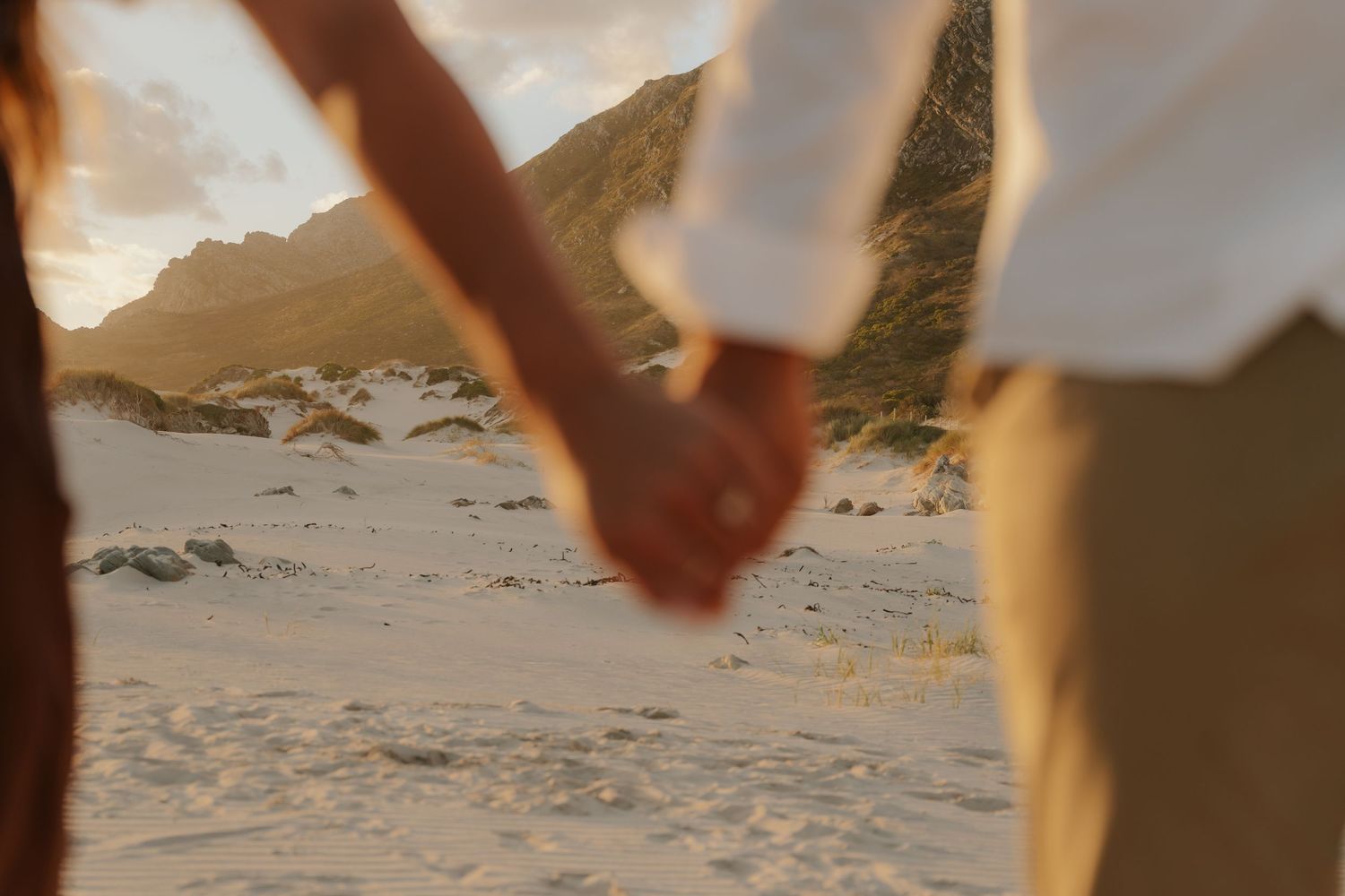 Couple holding hands on a sandy beach at sunset with mountains in the background.