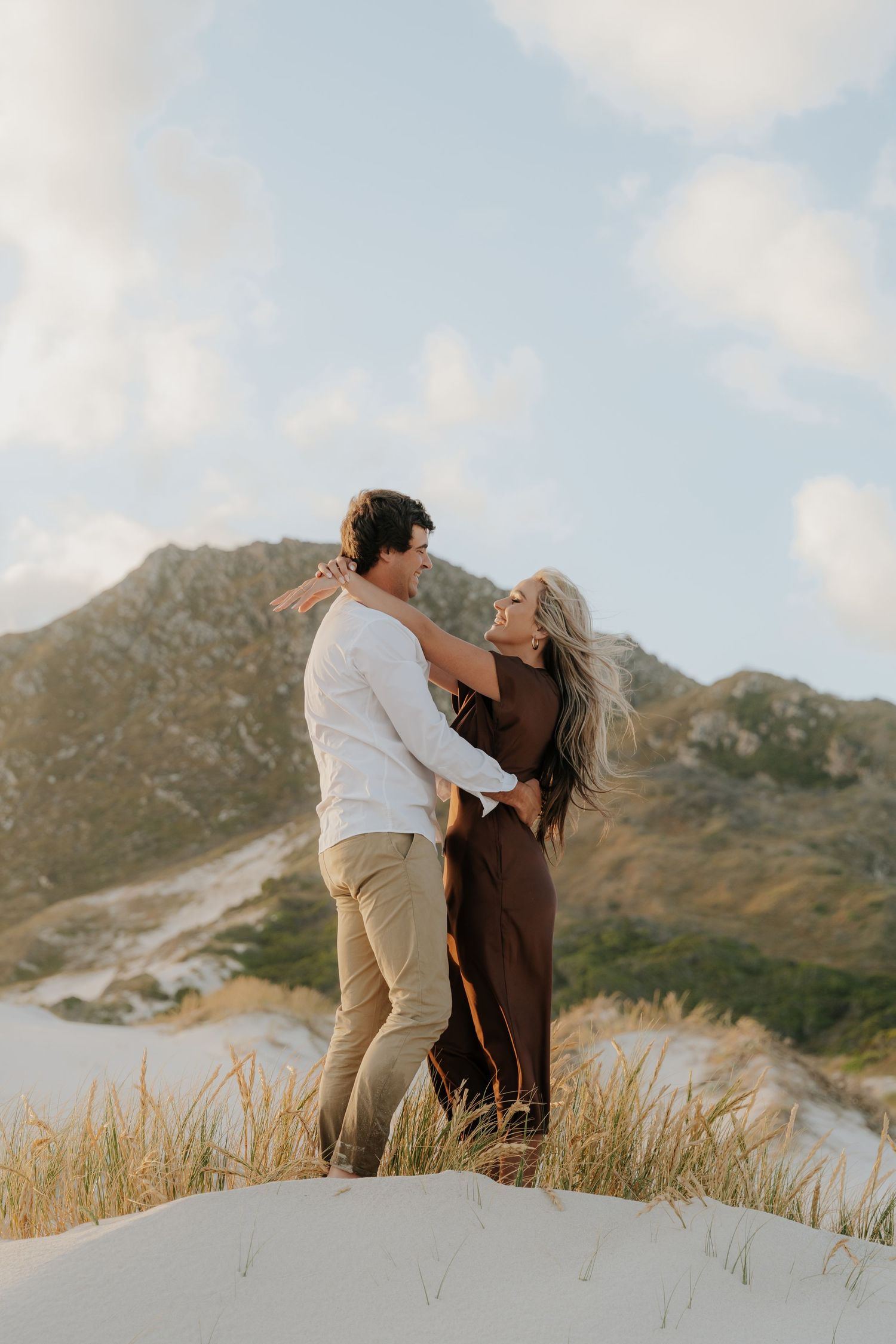 Couple embracing on sandy dunes with mountain backdrop, woman in brown dress, man in white shirt, golden hour light.