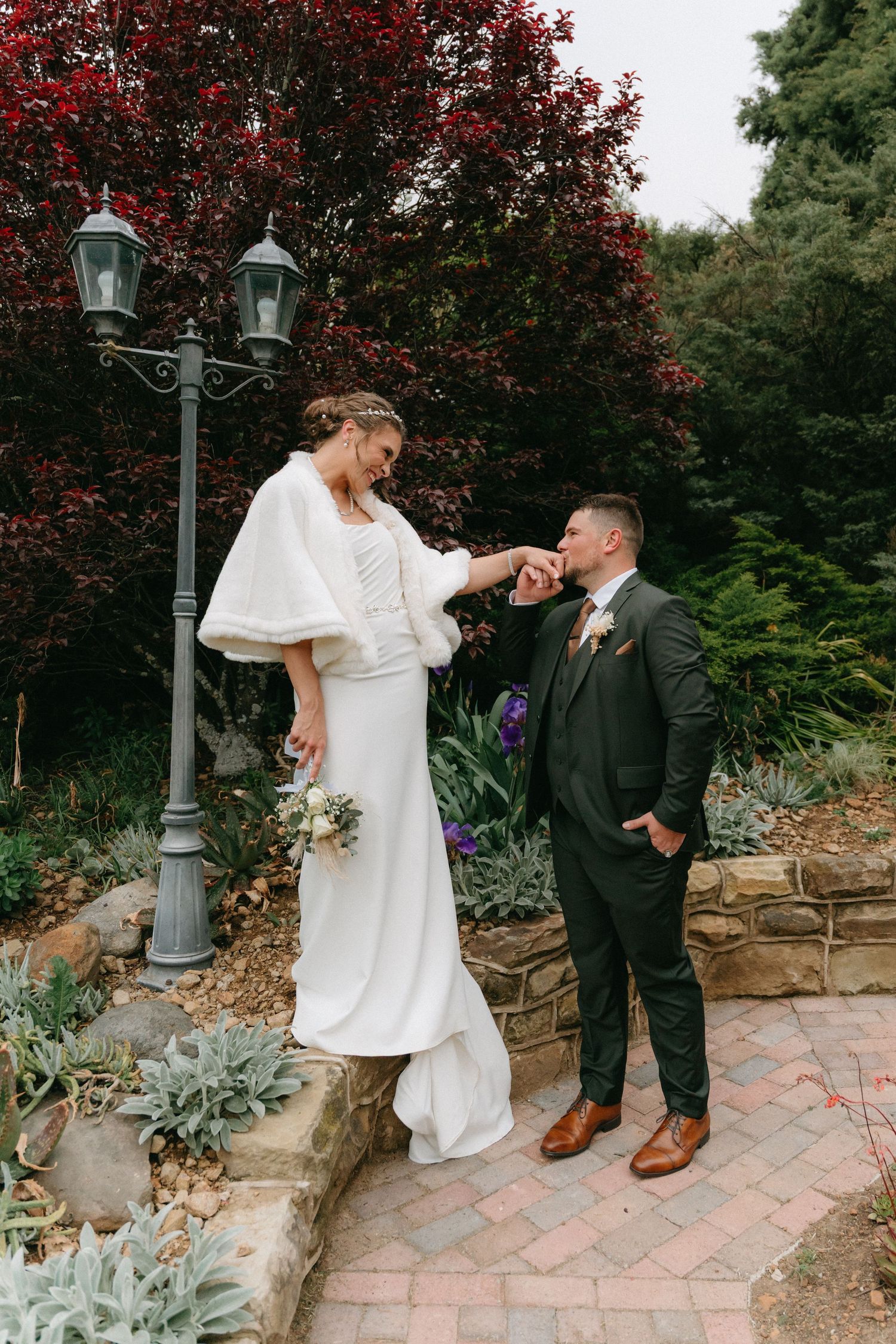 Wedding couple poses playfully on a brick path with red foliage and lamp posts.