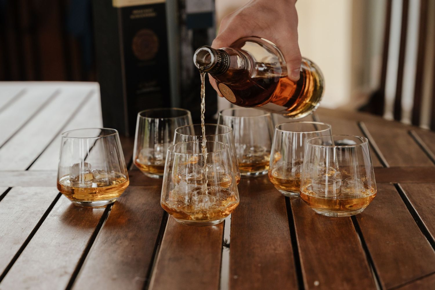 Multiple glasses of whiskey or bourbon lined up on a rustic wooden table in warm lighting.