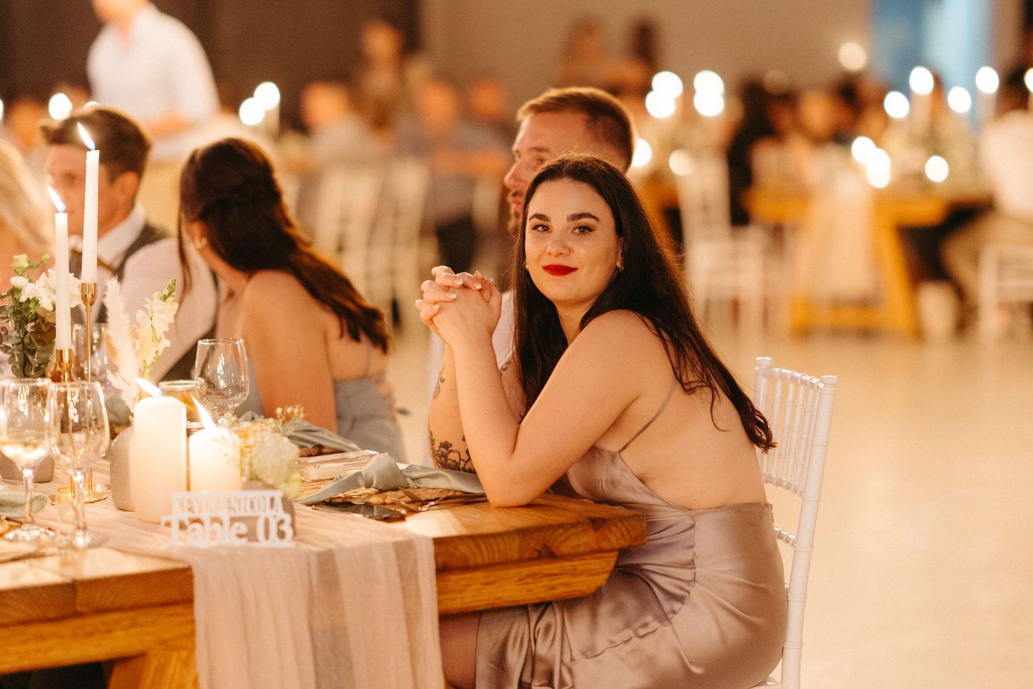 An elegant wedding guest in a silver dress sits at a beautifully decorated reception table with candlelight and warm ambiance.