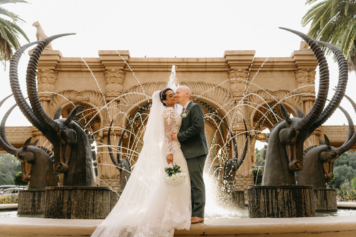 Newlyweds share a romantic moment under decorative elephant statues at an ornate outdoor venue.