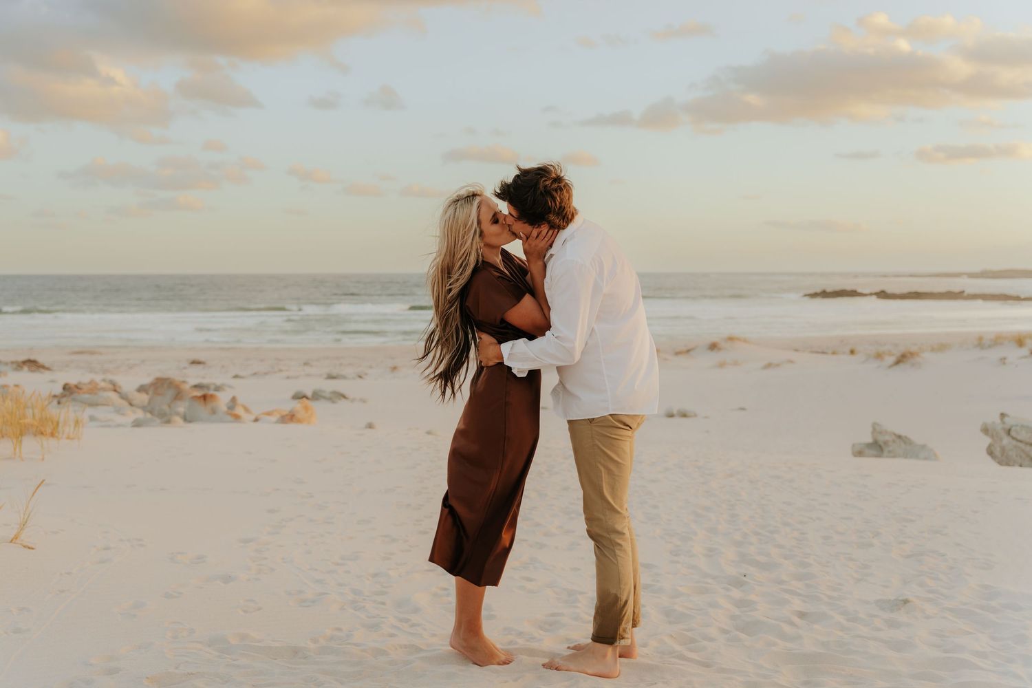 Couple kissing barefoot on white sand beach near Cape Town at sunset during romantic engagement photography session.