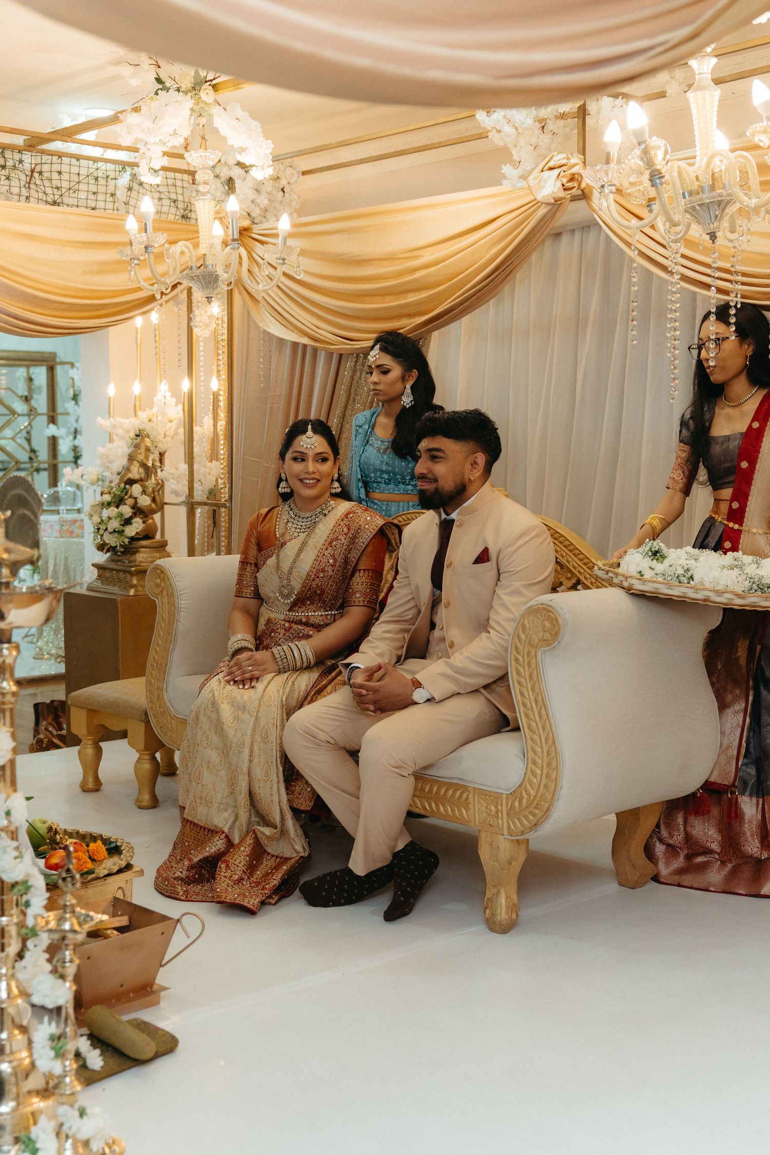 Couple in traditional South Asian wedding attire sits on ornate white chairs under elegant draped fabric and chandeliers.