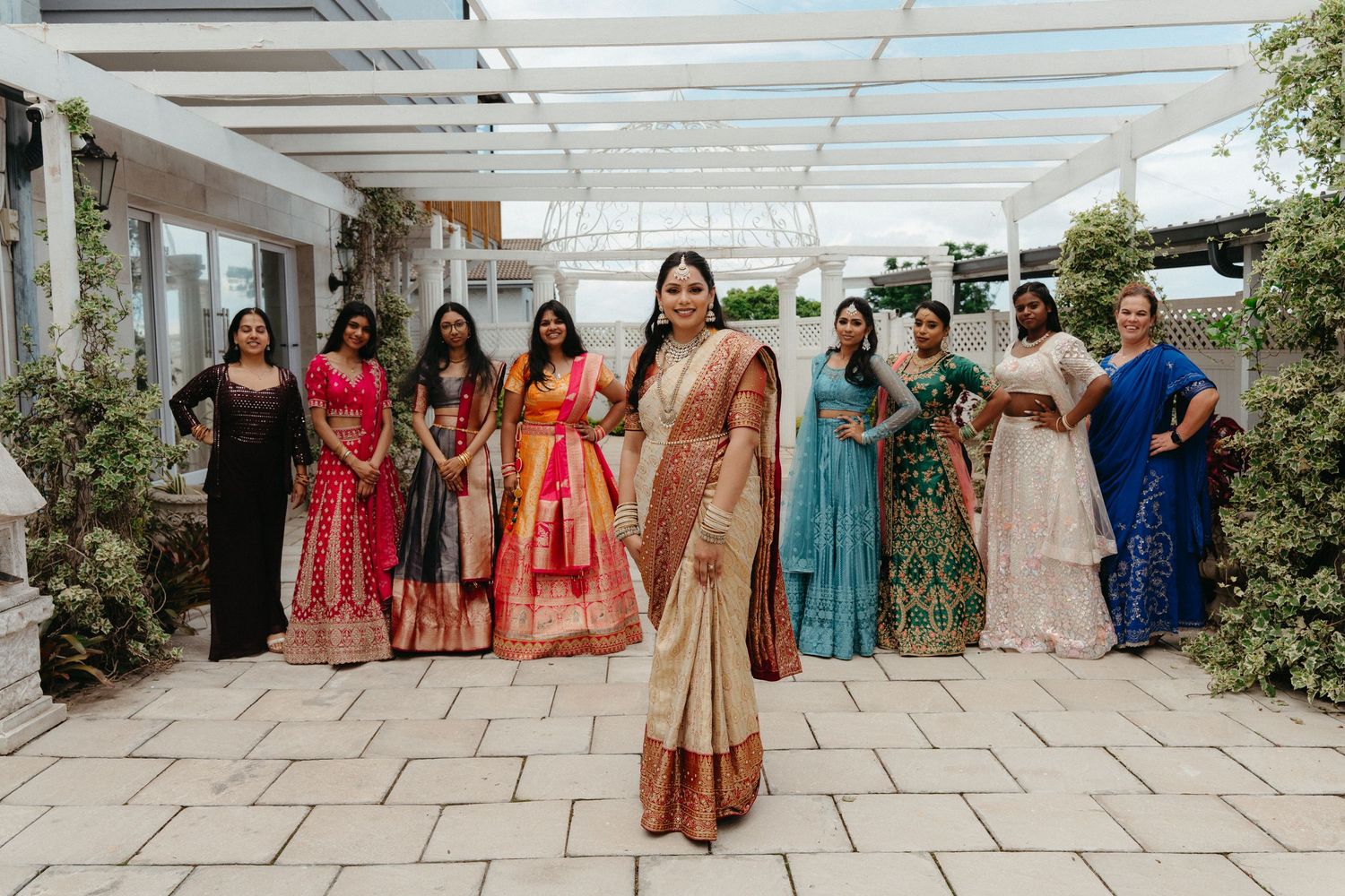 A group of people in traditional Indian attire pose together in a bright outdoor setting with white columns and greenery.