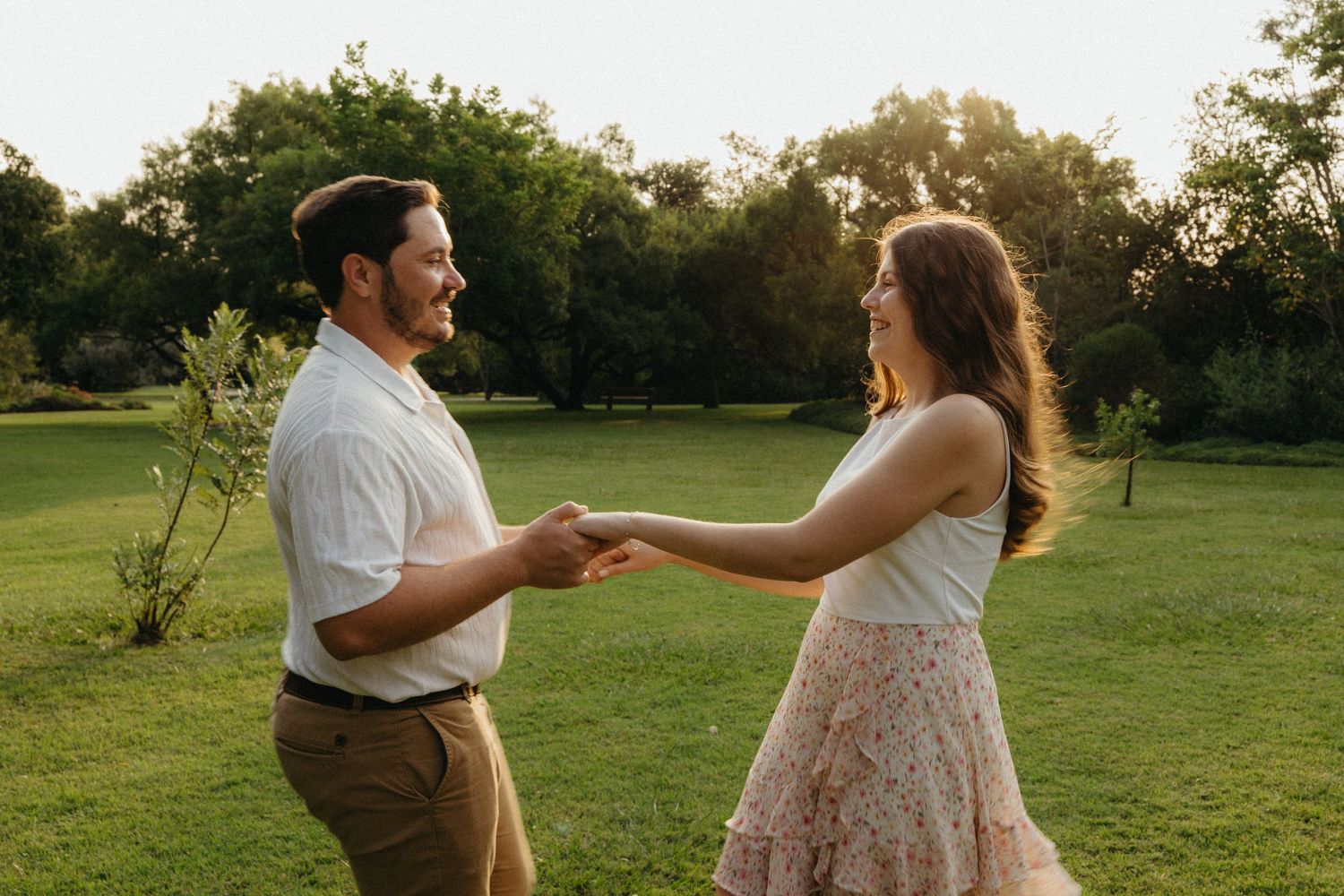 Couple holding hands and dancing together in a lush green park at golden hour sunset.