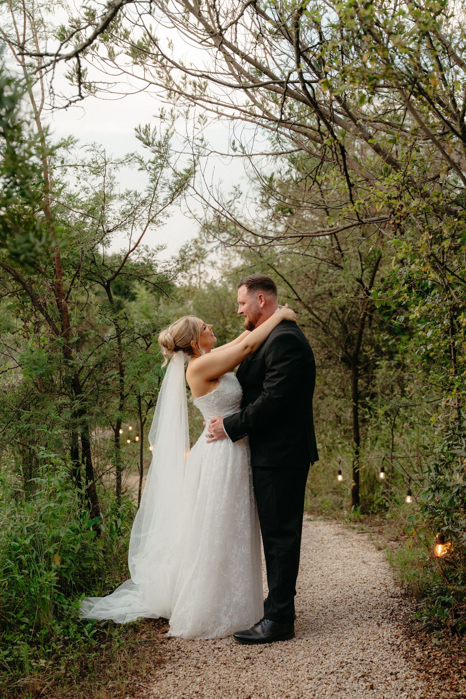 A romantic wedding couple shares an intimate moment on a tree-lined gravel path at sunset.