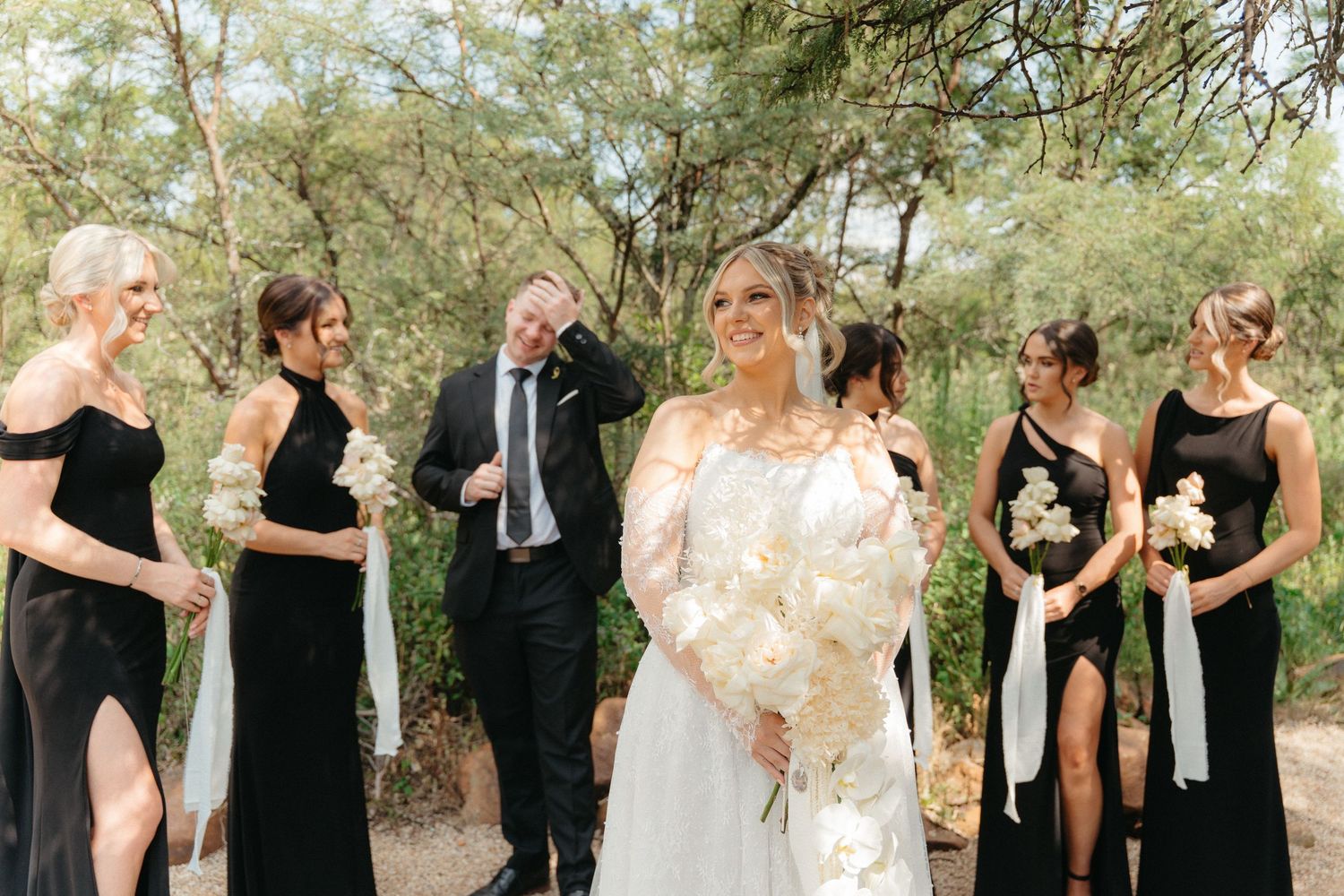 Wedding party in black and white attire poses together in an outdoor setting with trees in the background.
