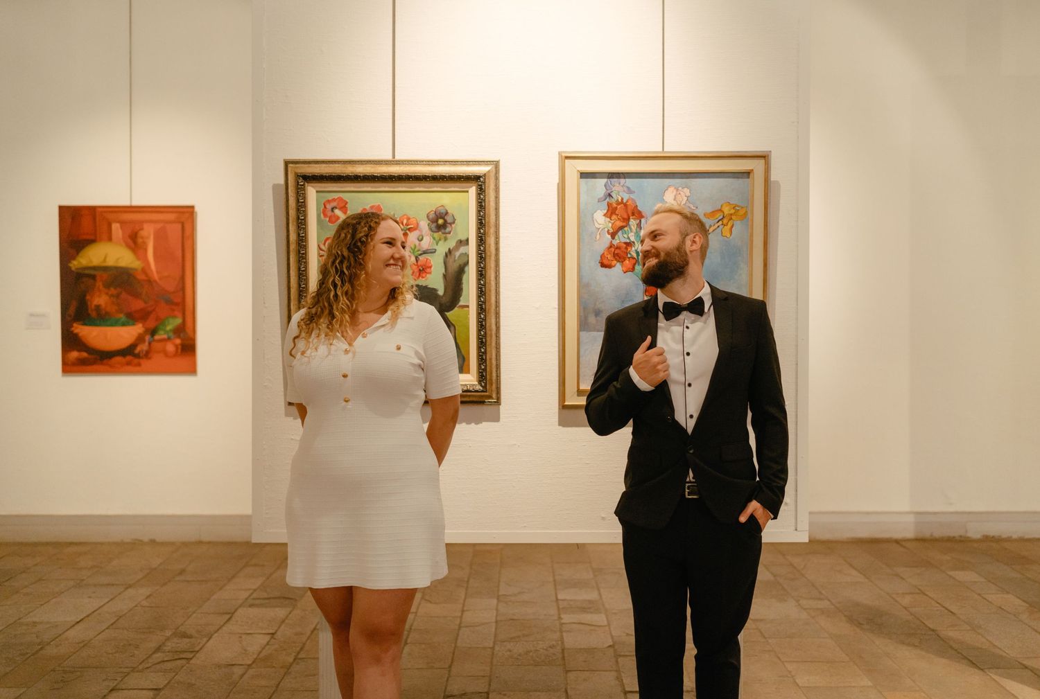Couple in formal attire sharing a lighthearted moment in art gallery with paintings.