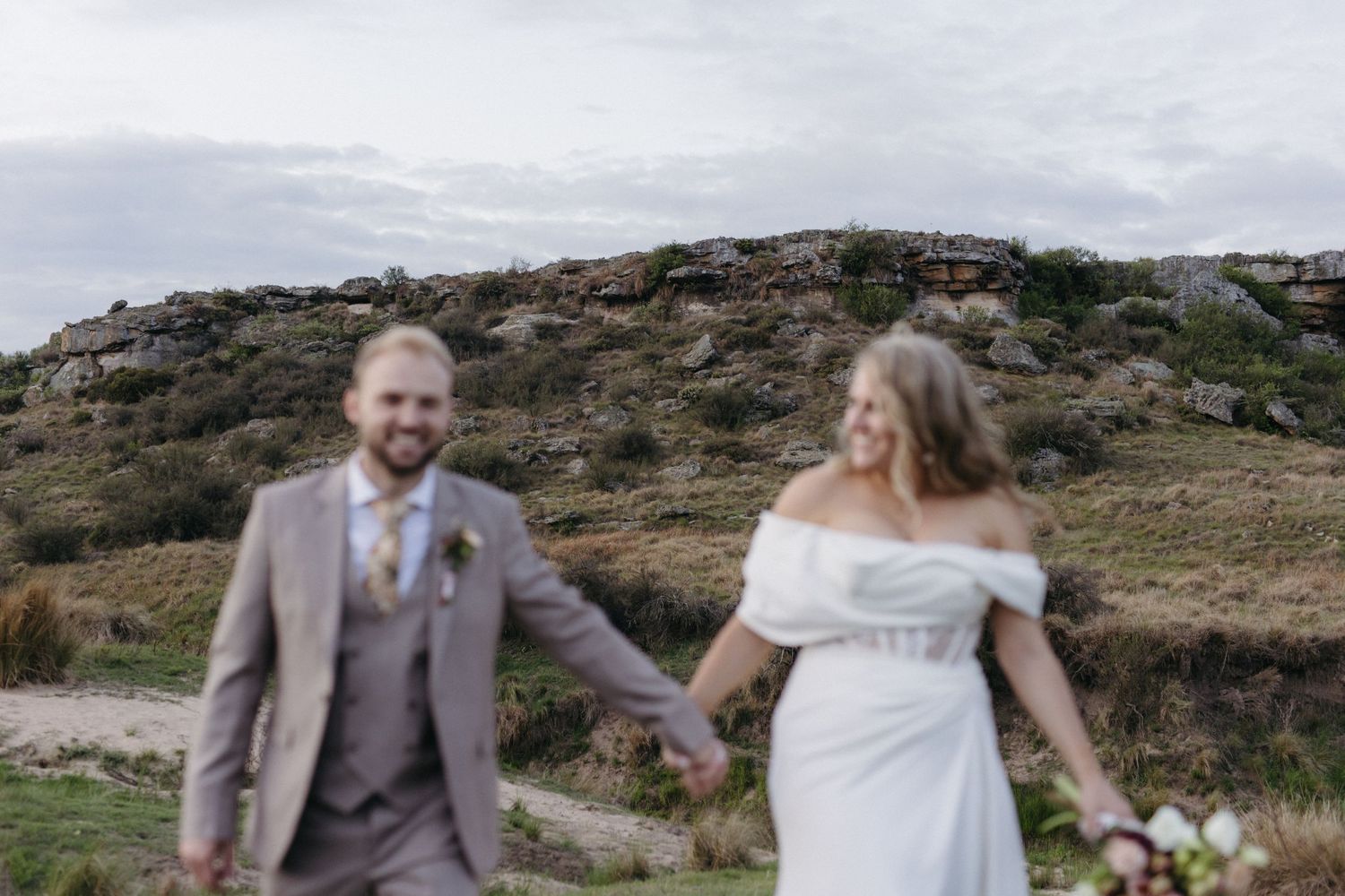 A couple in wedding attire hold hands while walking on a scenic hillside under cloudy skies.