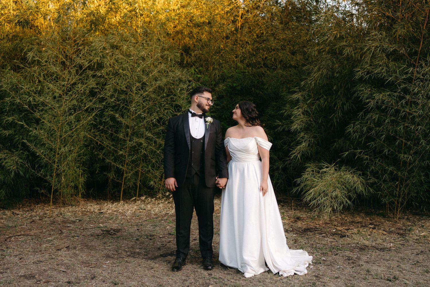 A couple in wedding attire stands together outdoors in front of golden bamboo during an autumn photoshoot.