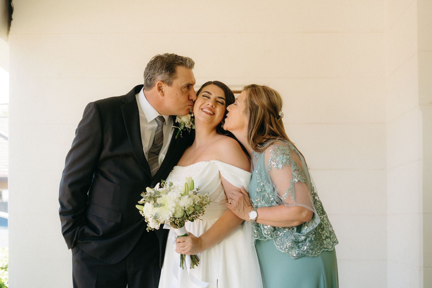 A bride in a white dress holds a bouquet of white flowers while receiving affectionate gestures at a wedding ceremony.
