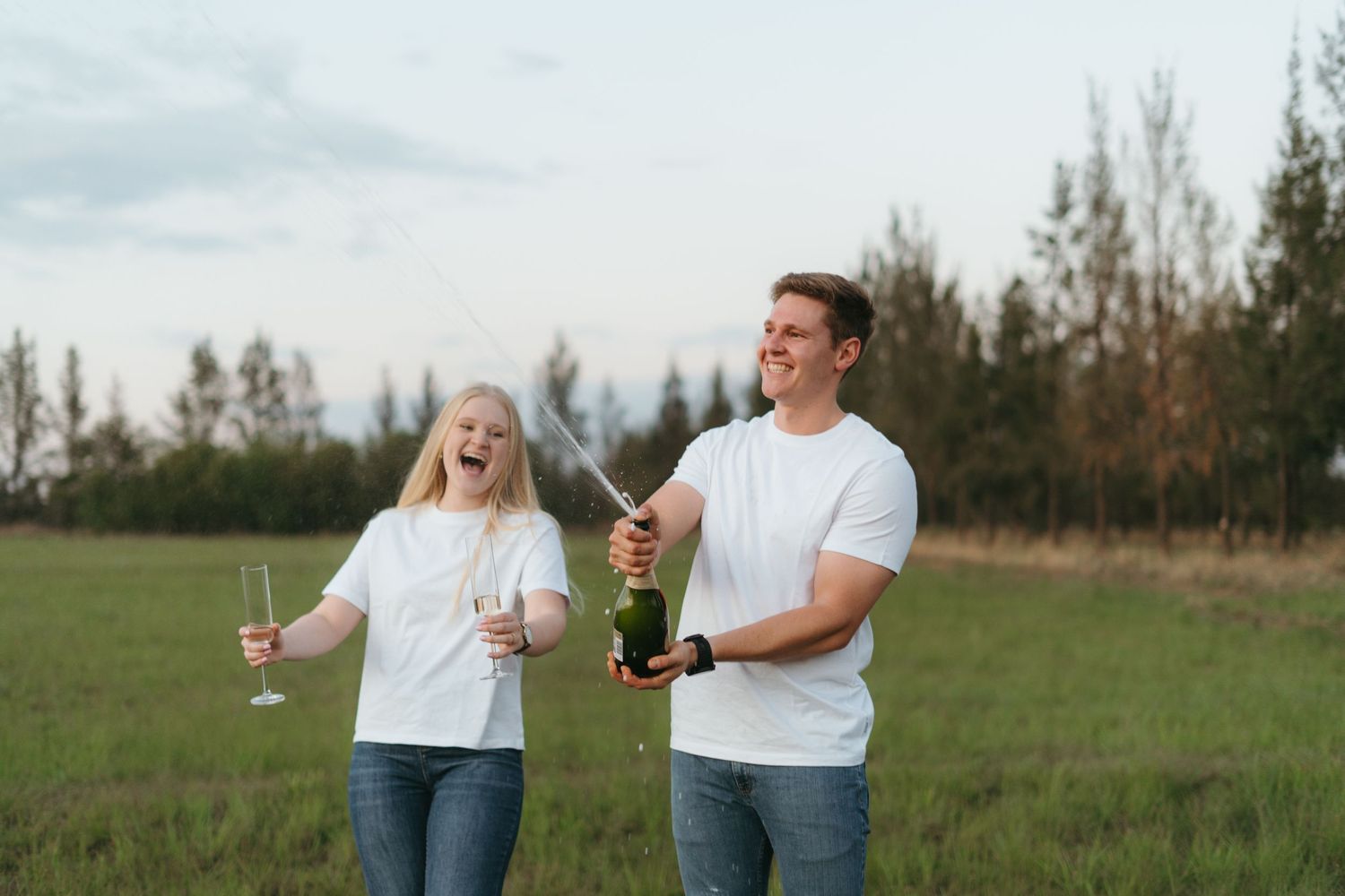 Couple laughing while spraying champagne in a field at sunset.