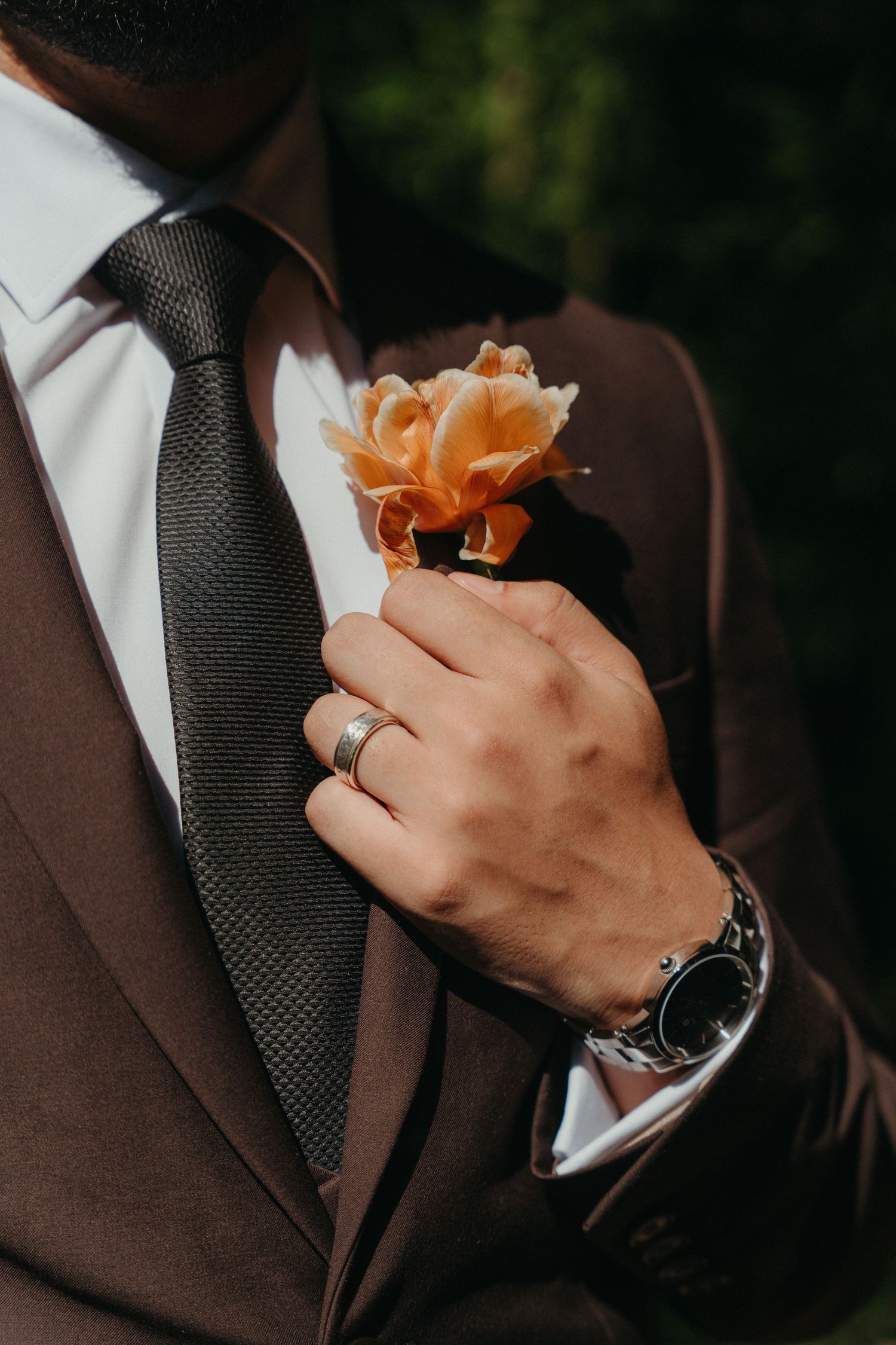 Groom in brown suit adjusting orange rose boutonniere while wearing wedding ring and silver watch at Beechwood Gardens, Johannesburg