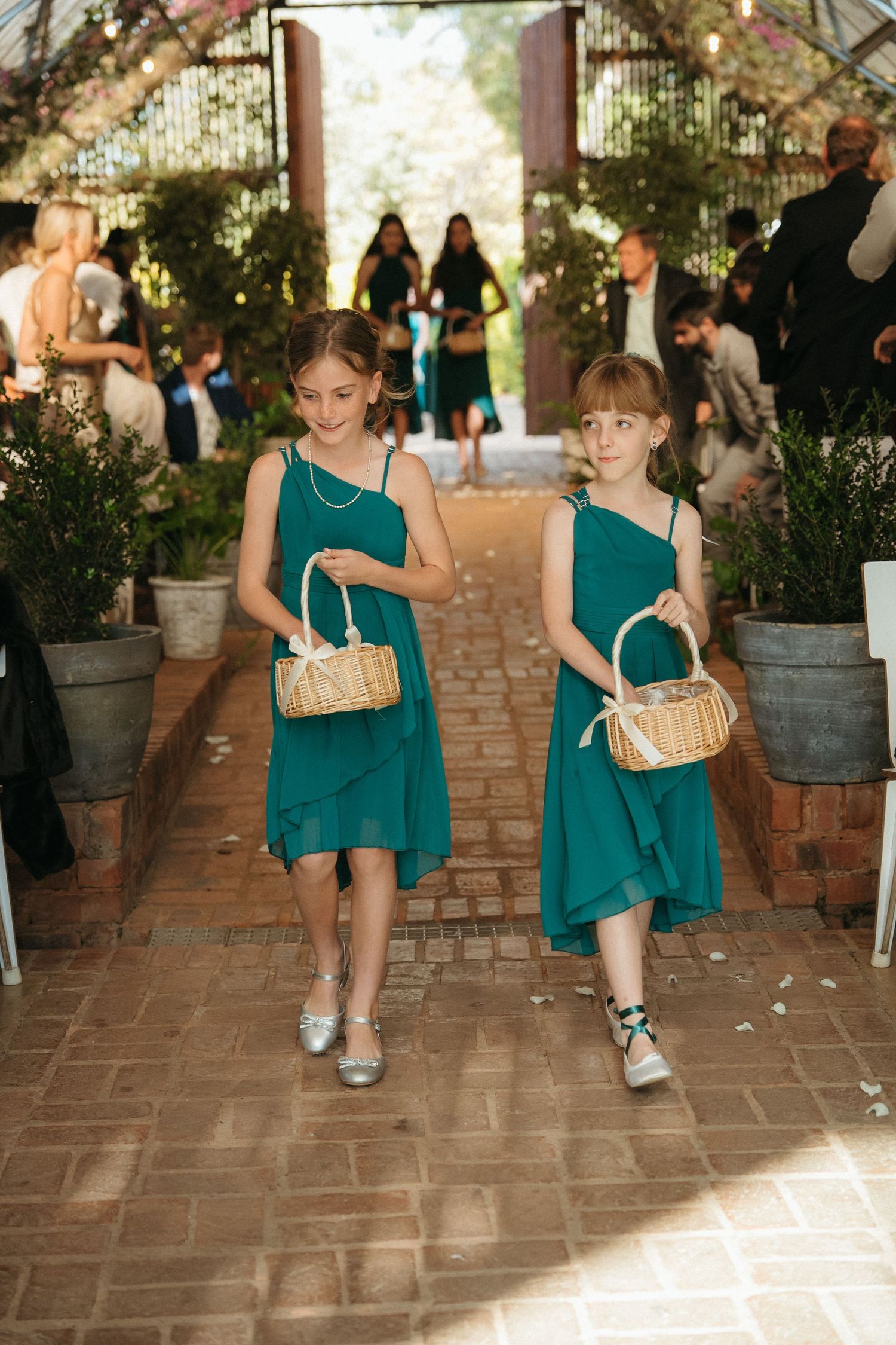 Two flower girls in teal dresses carrying wicker baskets walk down brick aisle at outdoor wedding ceremony.