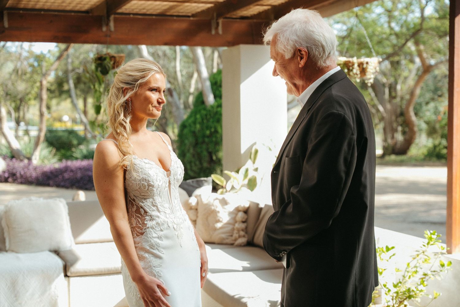 Bride in lace wedding gown shares a tender moment with an older gentleman in a black tuxedo at an outdoor venue.