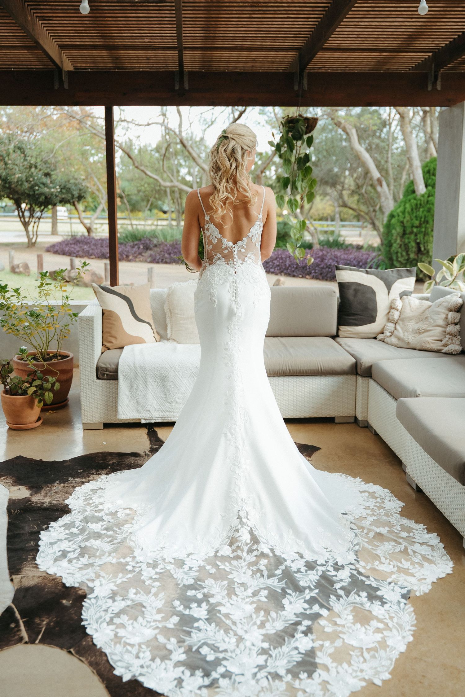 Bride in elegant backless lace mermaid wedding gown with floral train, standing in a sunlit outdoor venue.