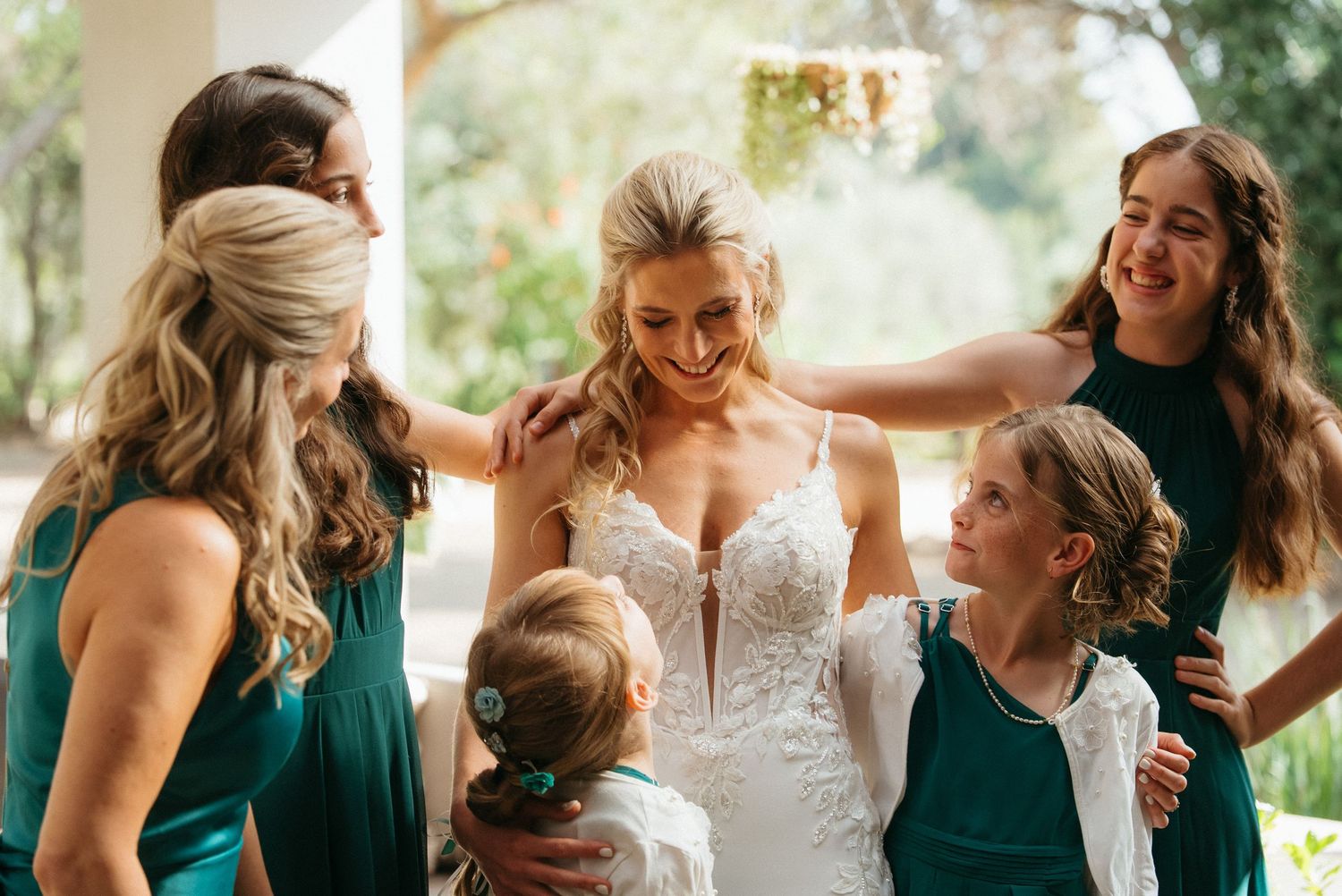 Bride in white lace gown smiles with bridesmaids and flower girl in teal dresses during wedding preparations.