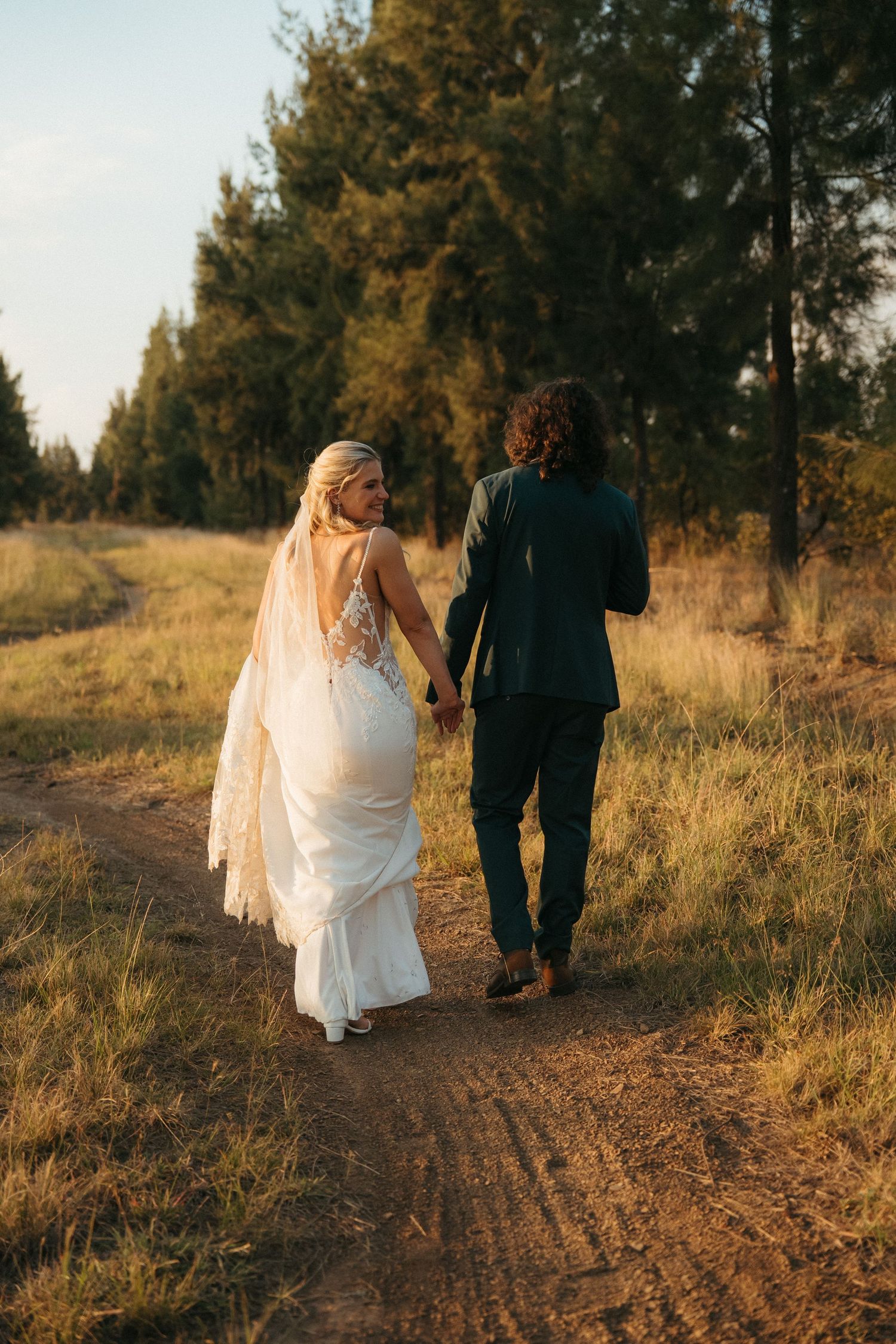 A bride and groom walk hand in hand down a rustic dirt path surrounded by golden grass and tall pine trees at sunset.