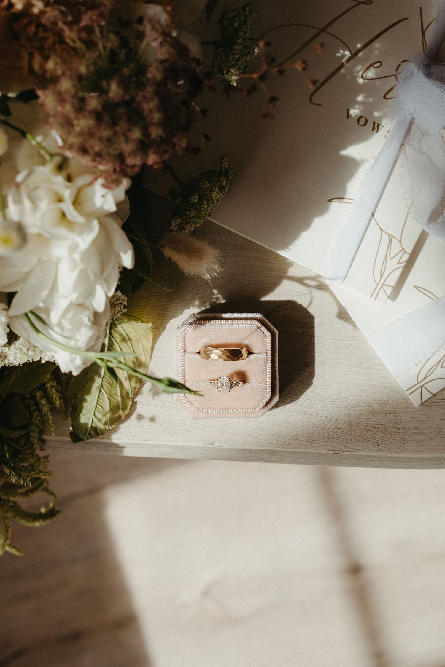 Gold wedding rings in a pink velvet box beside white flowers and wedding stationery in soft natural light.