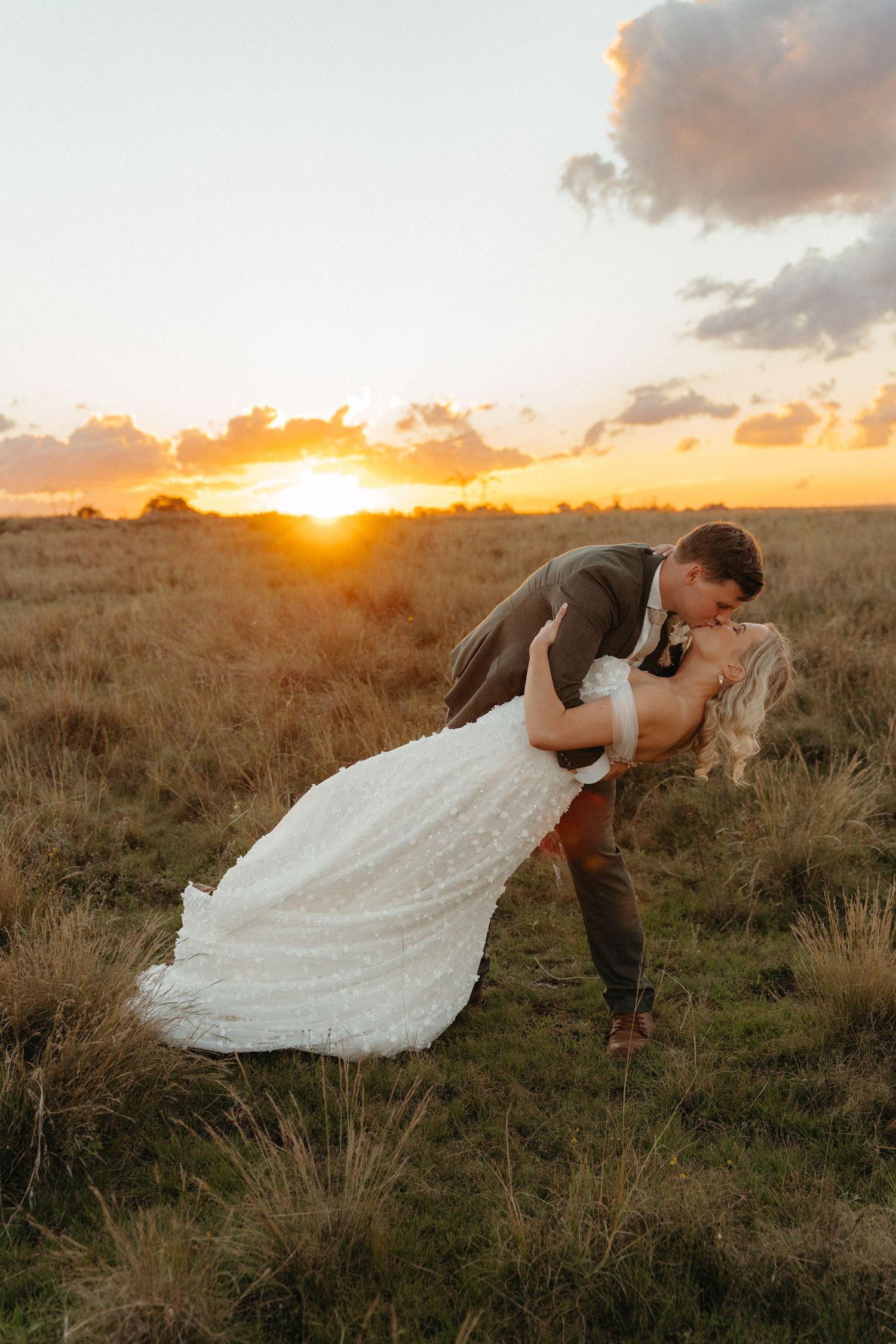 Bride and groom share a romantic dip kiss at golden sunset in an open field, bride in white lace gown.