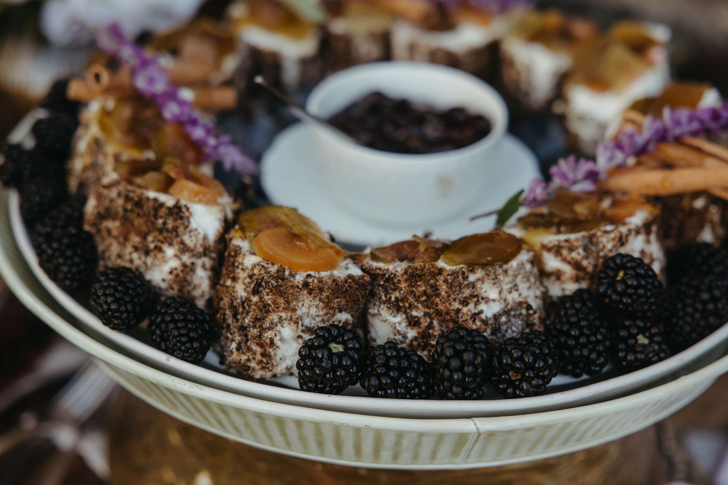 Elegant dessert platter with blackberries, chocolate-dusted cake, cinnamon sticks, purple flowers, and a bowl of jam.