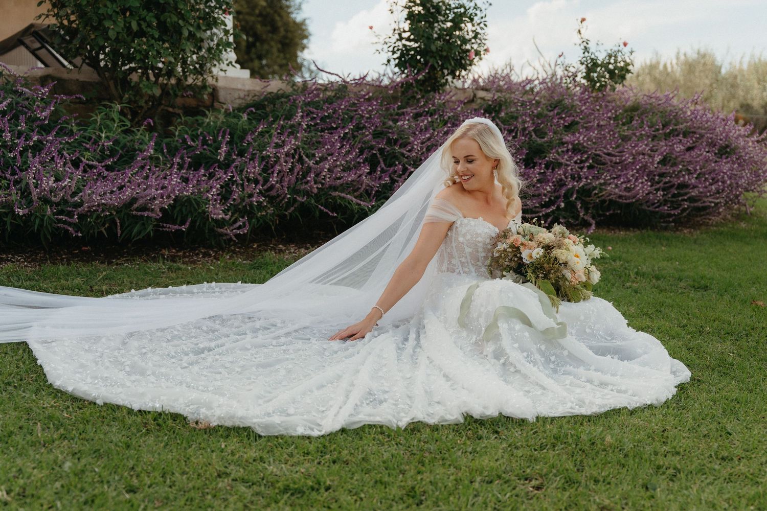 Bride in lace ball gown wedding dress with veil sits on green grass surrounded by purple flowering bushes holding bouquet.