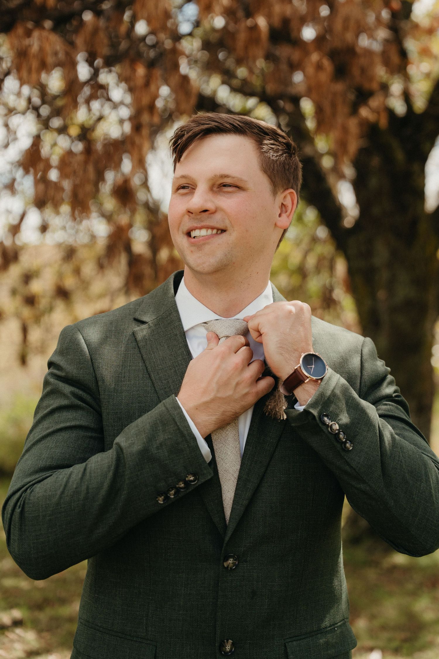 Smiling groom in green tweed suit adjusting his tie outdoors with autumn trees in background.