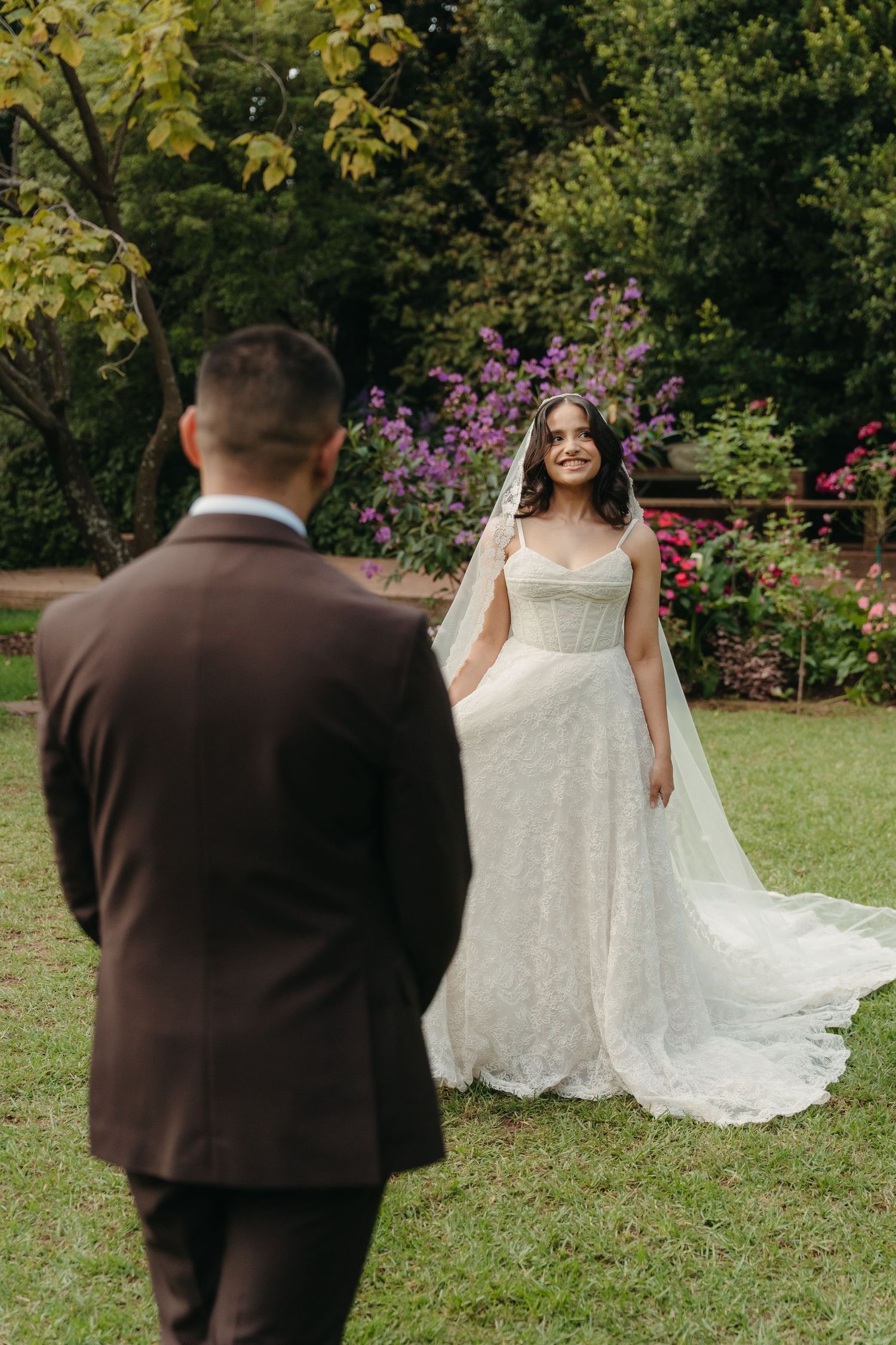 Bride in white lace gown and veil smiles at groom in brown suit during outdoor garden wedding ceremony.