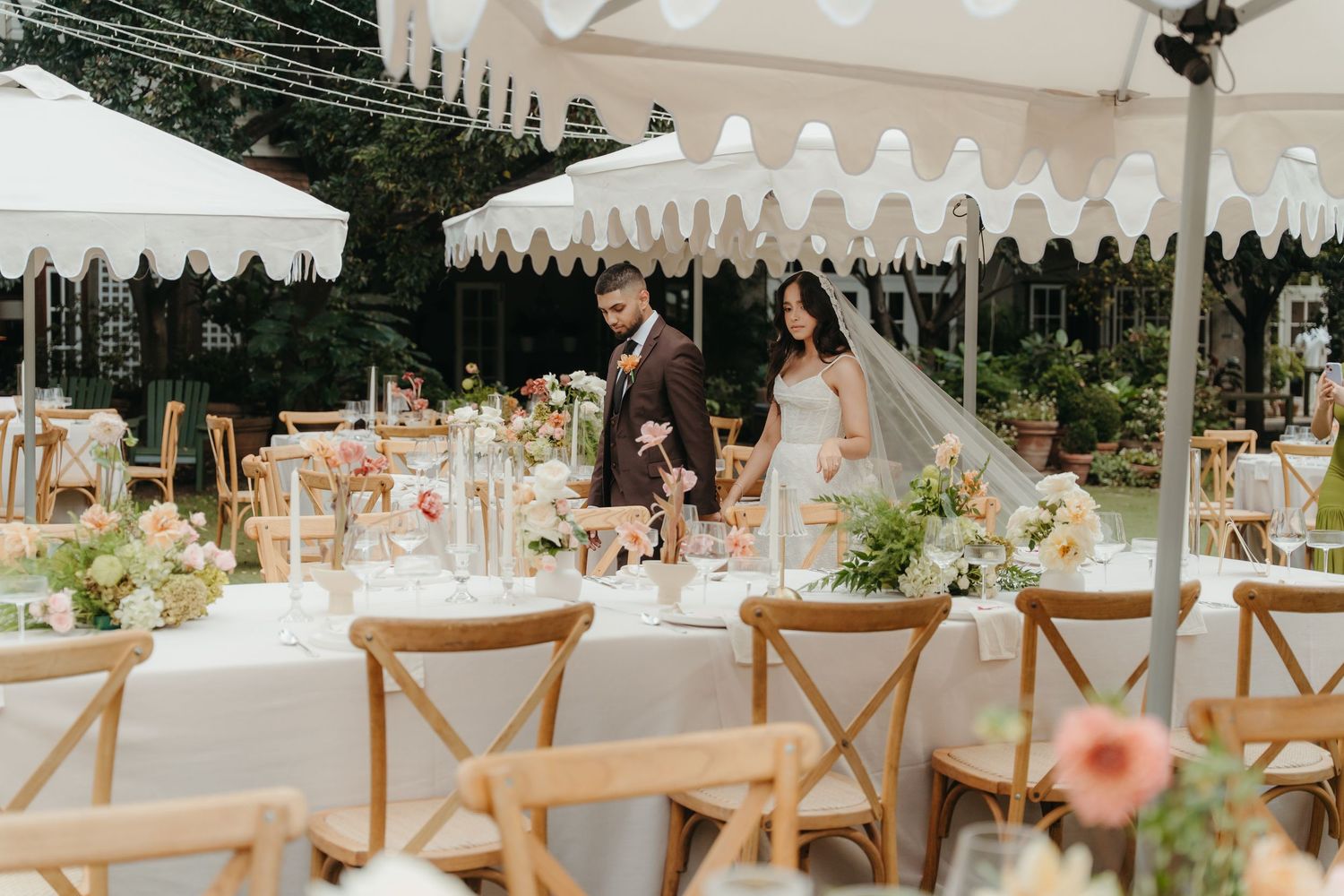 Bride and groom walk through an elegant outdoor wedding reception with floral centerpieces and white scalloped umbrellas.