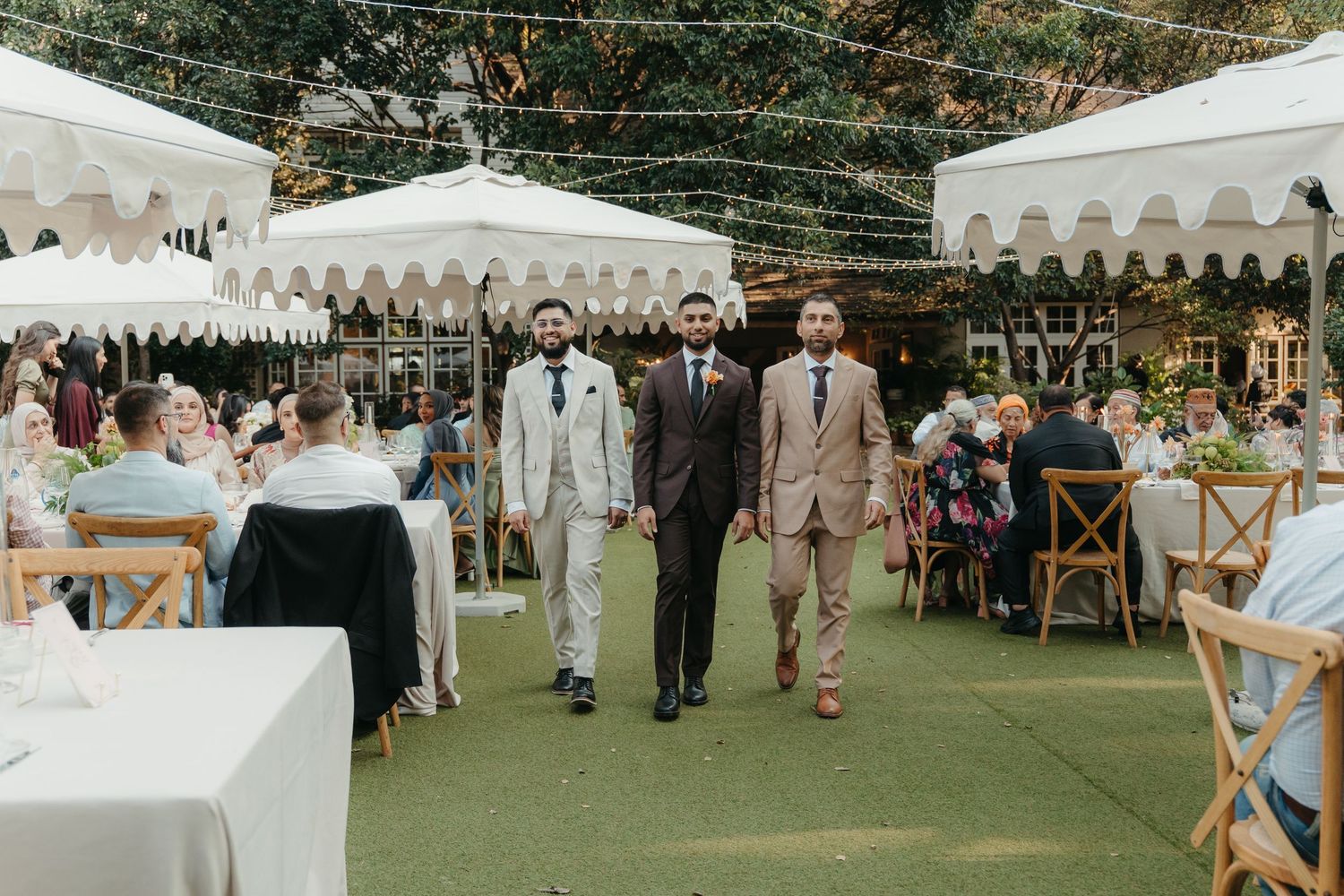 Three groomsmen in suits walk down outdoor wedding aisle with white tent canopies and string lights.