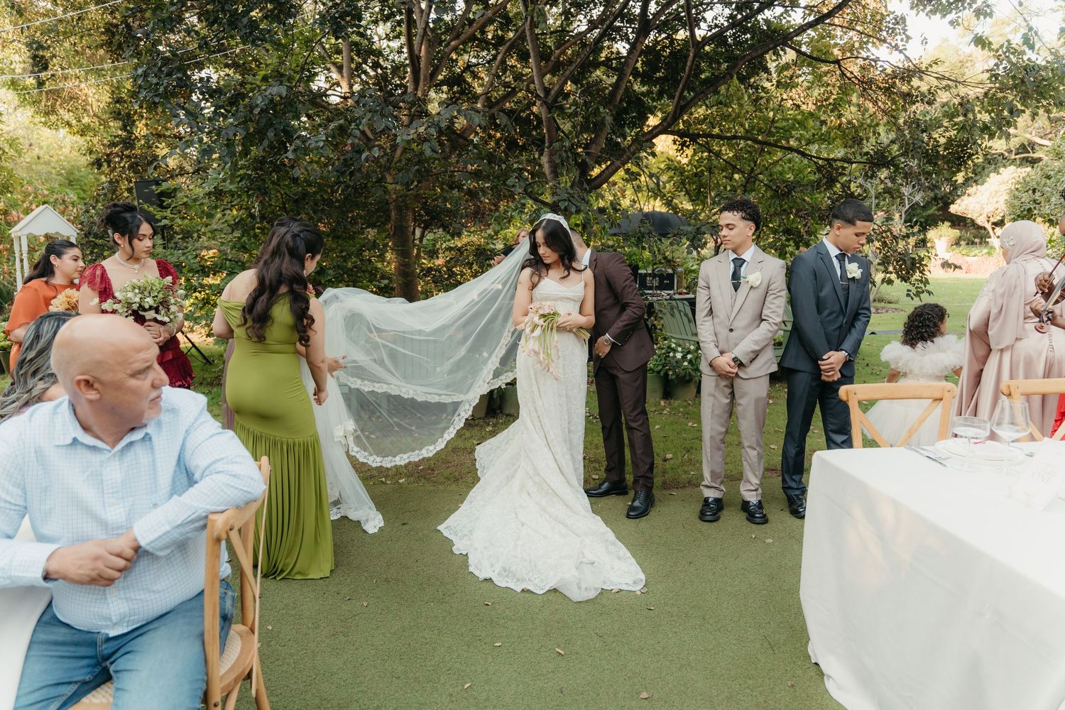 Outdoor wedding ceremony with bride in lace gown and veil, groom, and guests gathered on green lawn surrounded by trees.