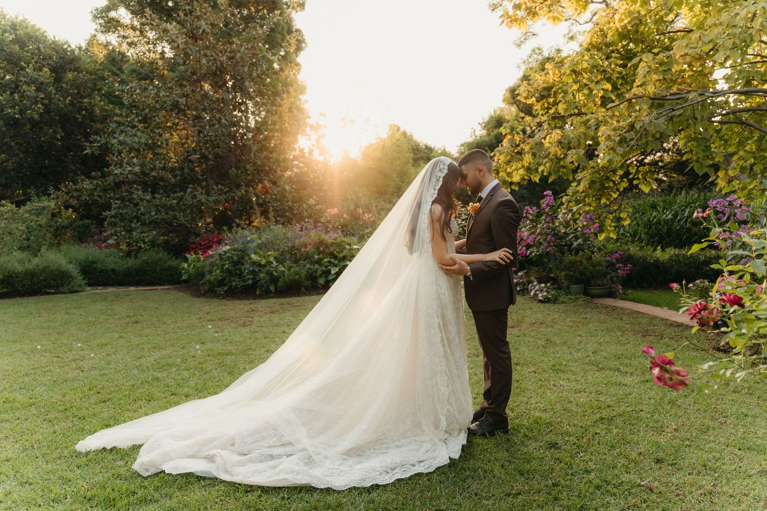 Bride and groom sharing a romantic kiss in a lush garden at golden hour, with a flowing cathedral veil.