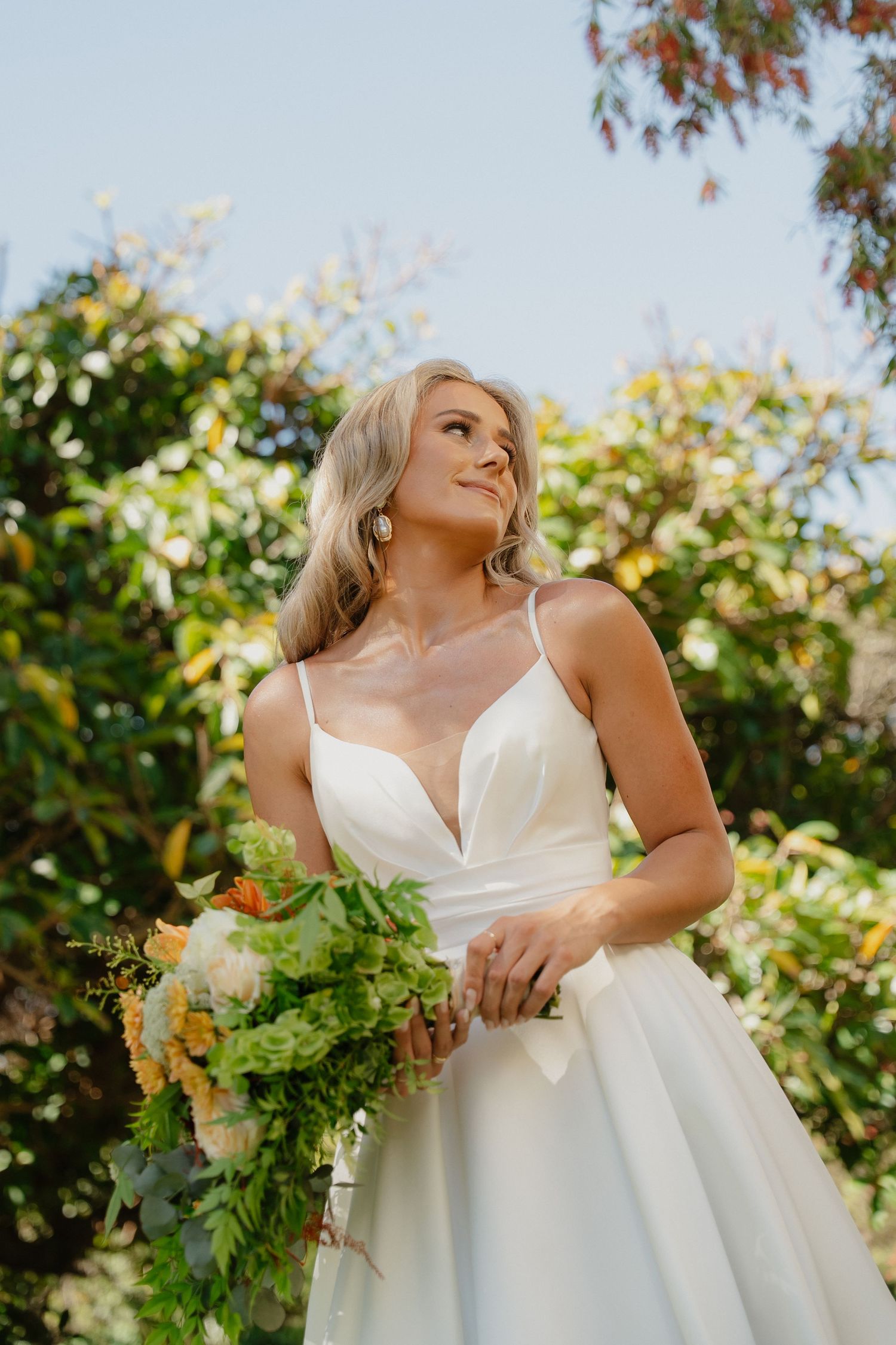 Blonde bride in white satin gown holding lush green bouquet outdoors, gazing upward with elegant drop earrings.
