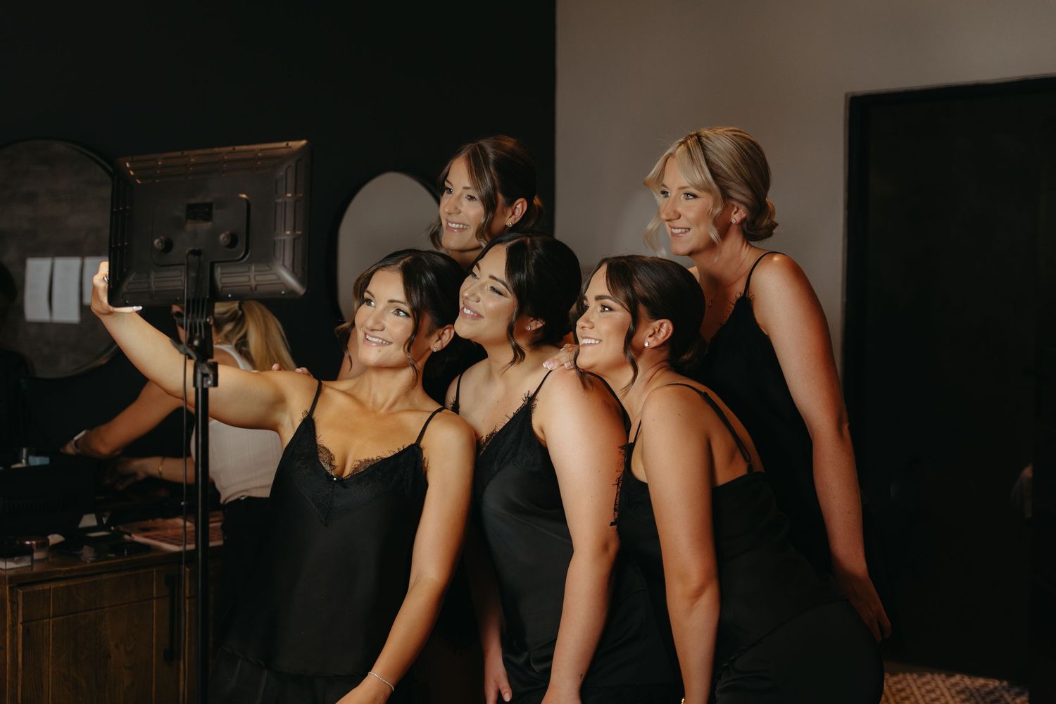 Wedding party members in dark dresses gather together in a dimly lit room during preparations.